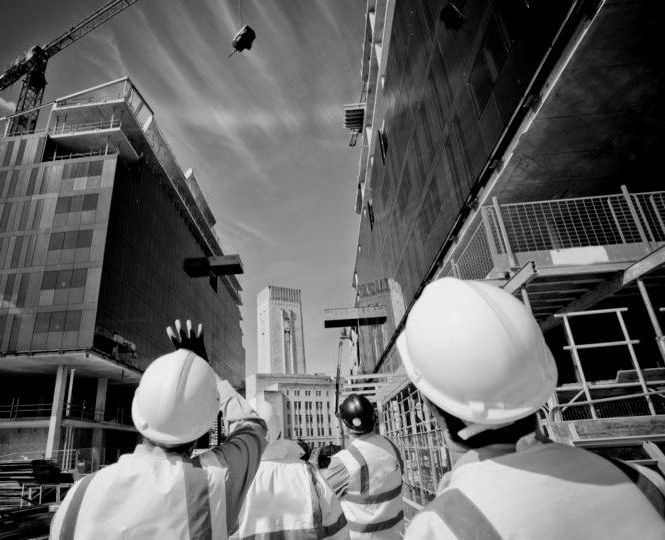 Trabalhadores da construção civil usando capacetes observam algo caindo em um canteiro de obras.