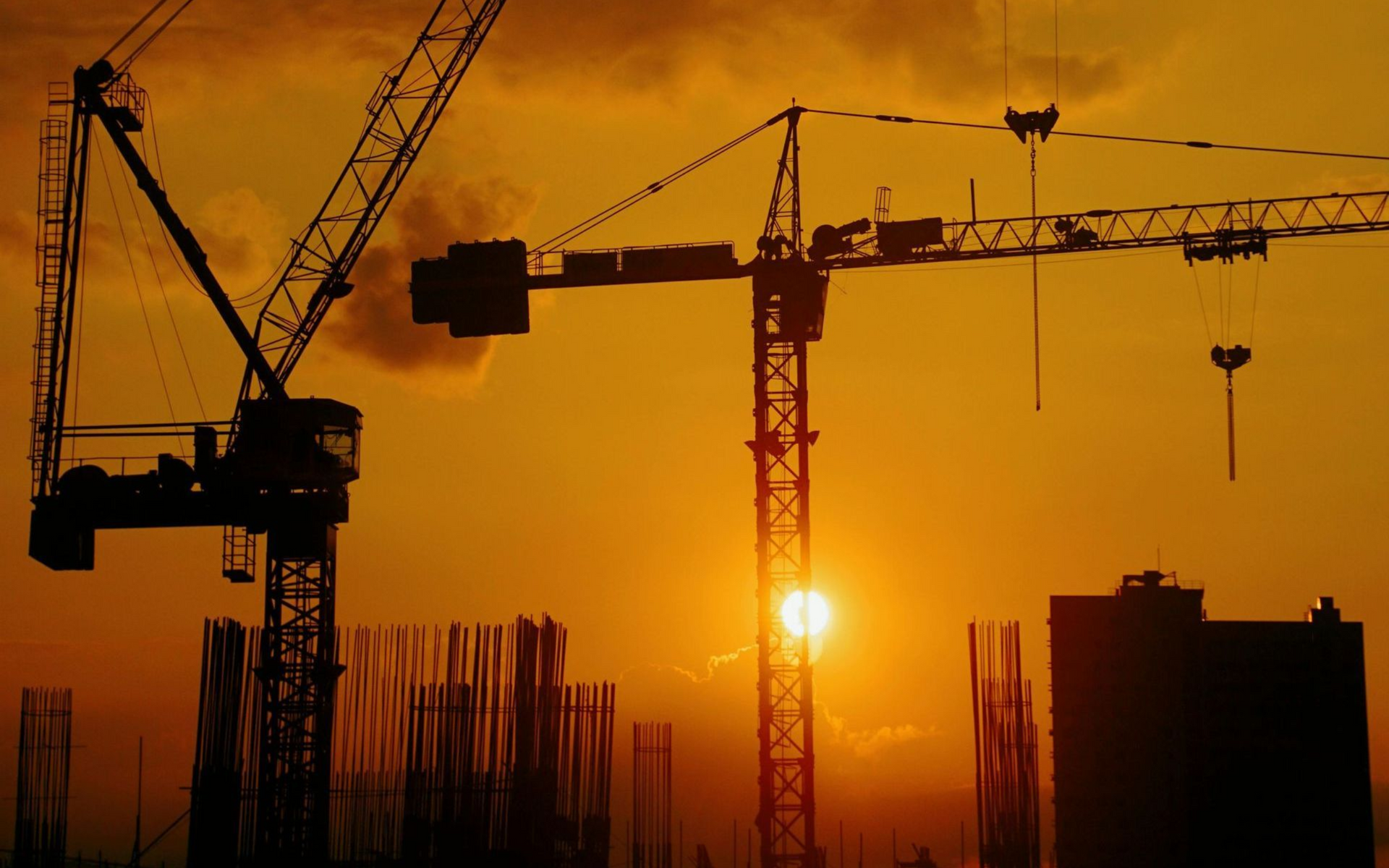 Guindastes de construção silhuetados contra um pôr do sol dourado sobre uma paisagem urbana, criando um cenário vibrante.