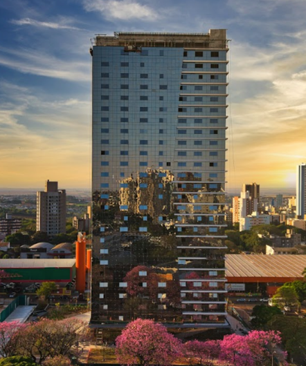 Prédio alto com fachada espelhada refletindo os arredores, em um cenário urbano ao pôr do sol, com árvores floridas cor-de-rosa.