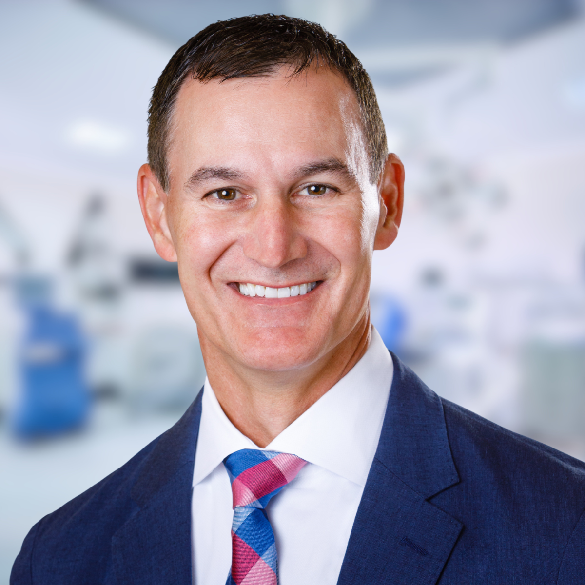 Smiling man in a blue suit and patterned tie, set against a blurred medical office background.