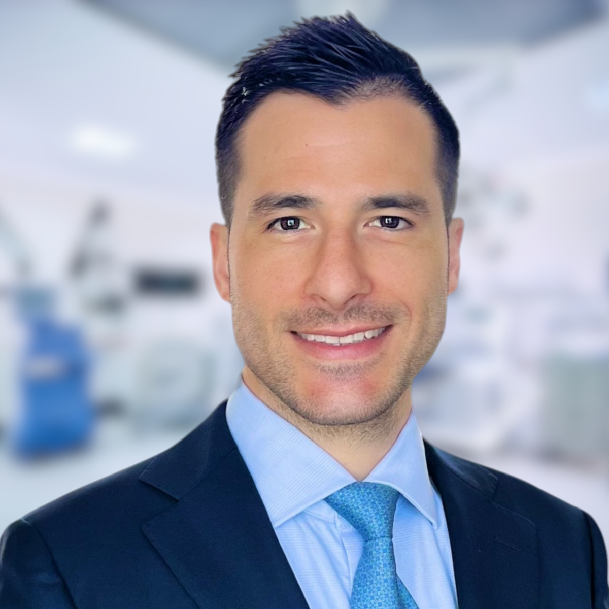 Man in a suit and tie smiling, set in a blurred medical office.