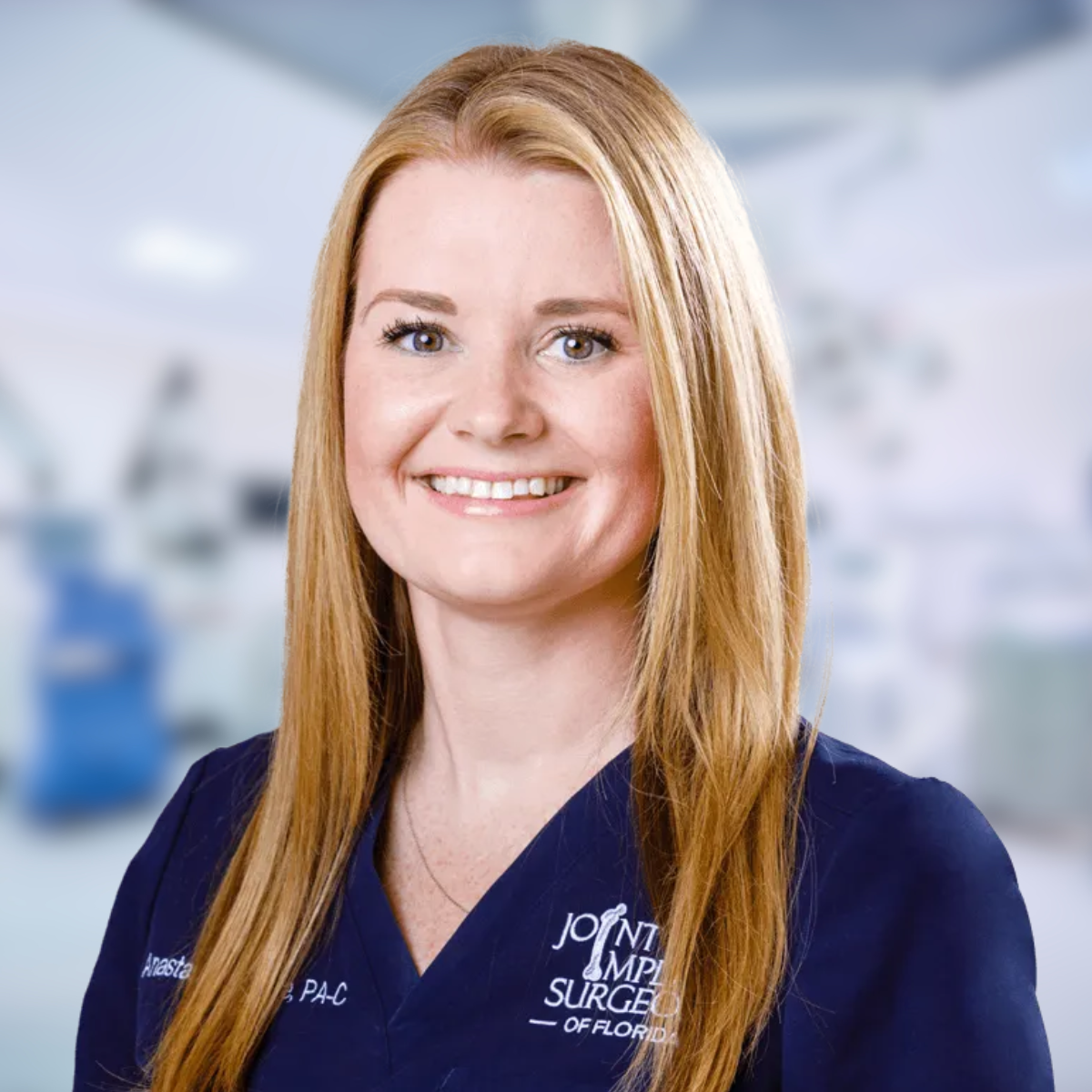 Woman with blonde hair in navy scrubs smiling, blurred surgical setting in background.