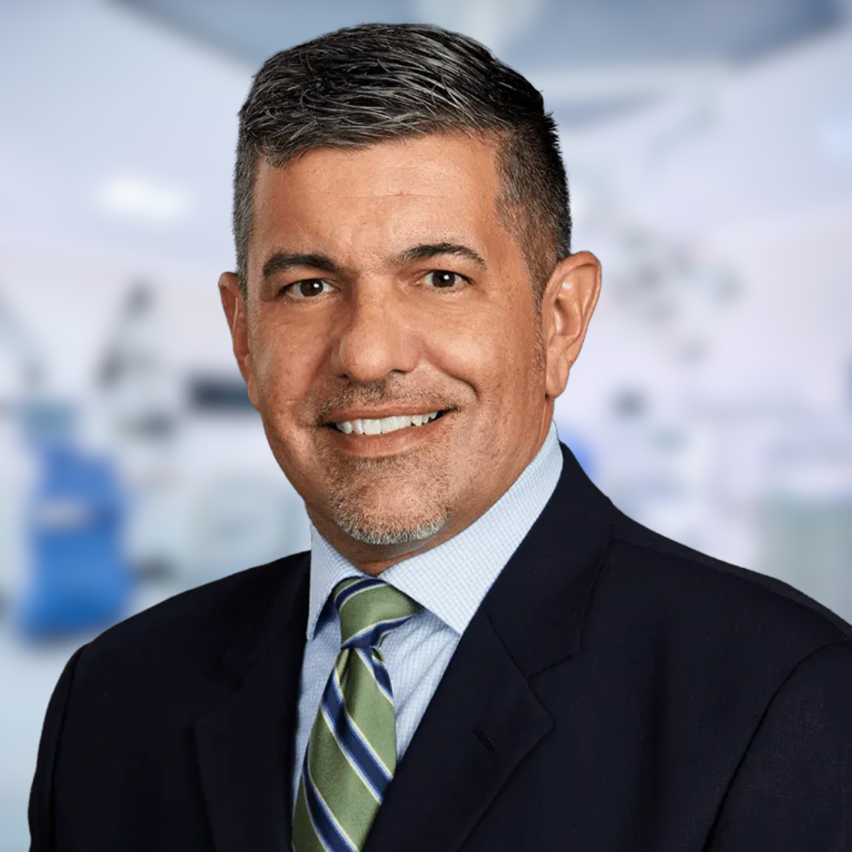Man in navy suit with green tie, smiling against blurred hospital background.