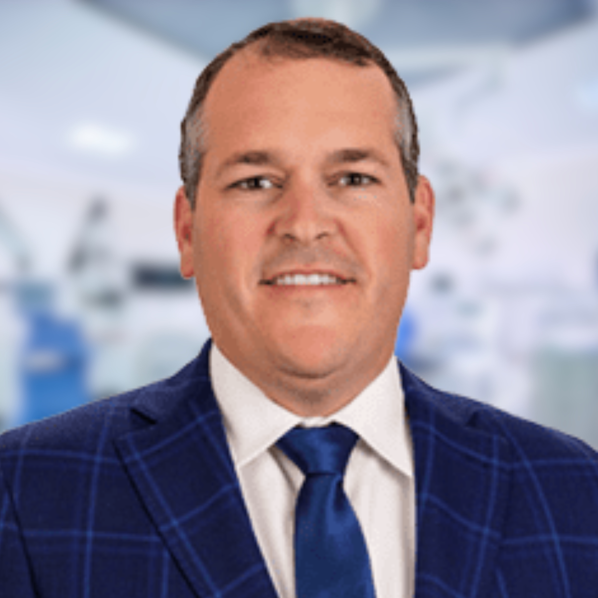Man in blue plaid suit and tie, smiling, set in an indistinct office environment.