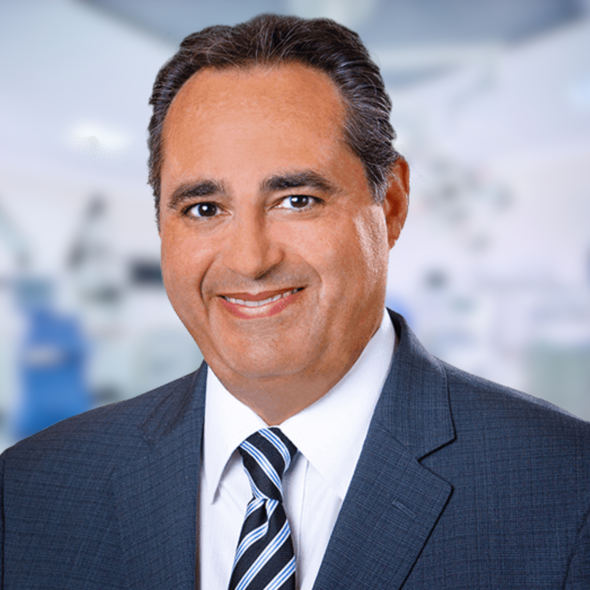 Smiling man in a suit and tie, possibly a doctor, in a blurred medical setting.