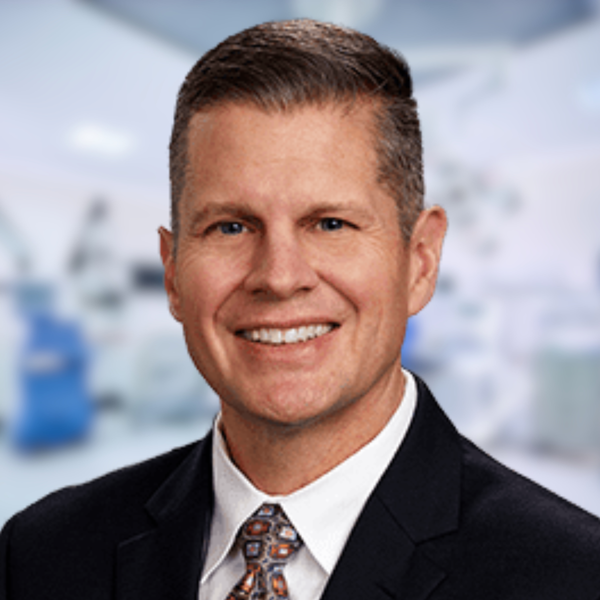 Man in suit smiling; blurred operating room background.