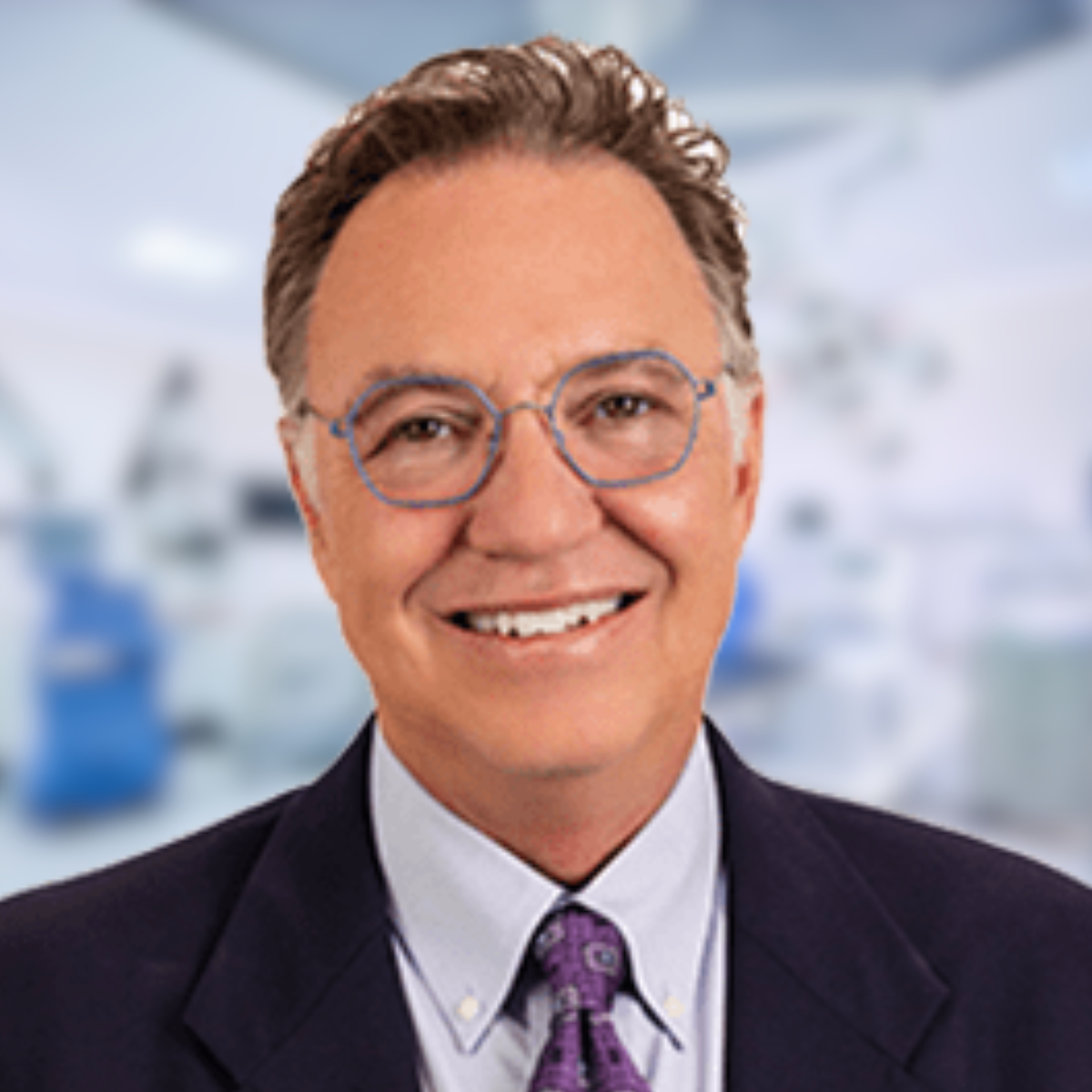 Smiling man in glasses, suit, and tie against a blurred medical setting.