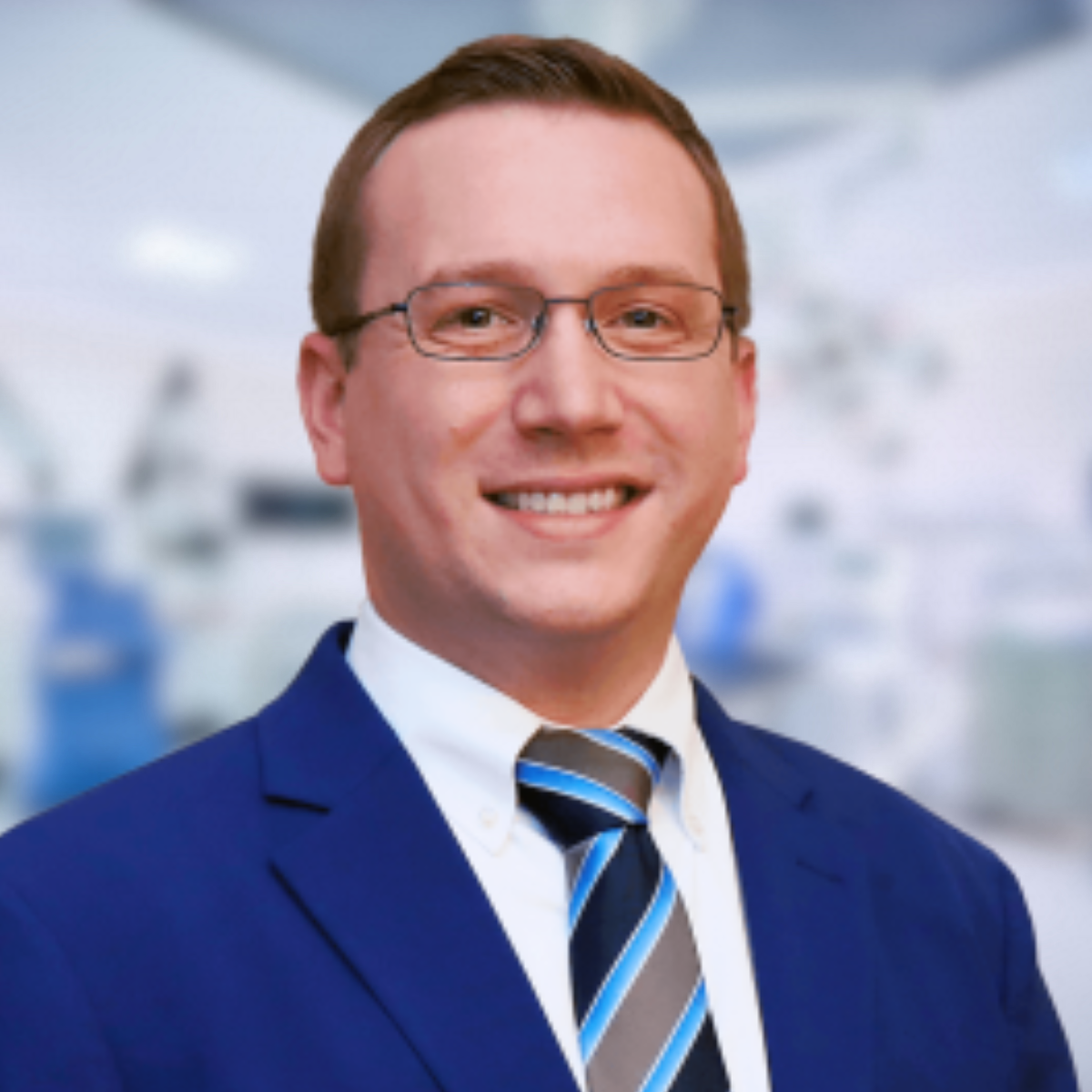 Man in blue suit and tie smiles at the camera, with glasses, in a medical setting.