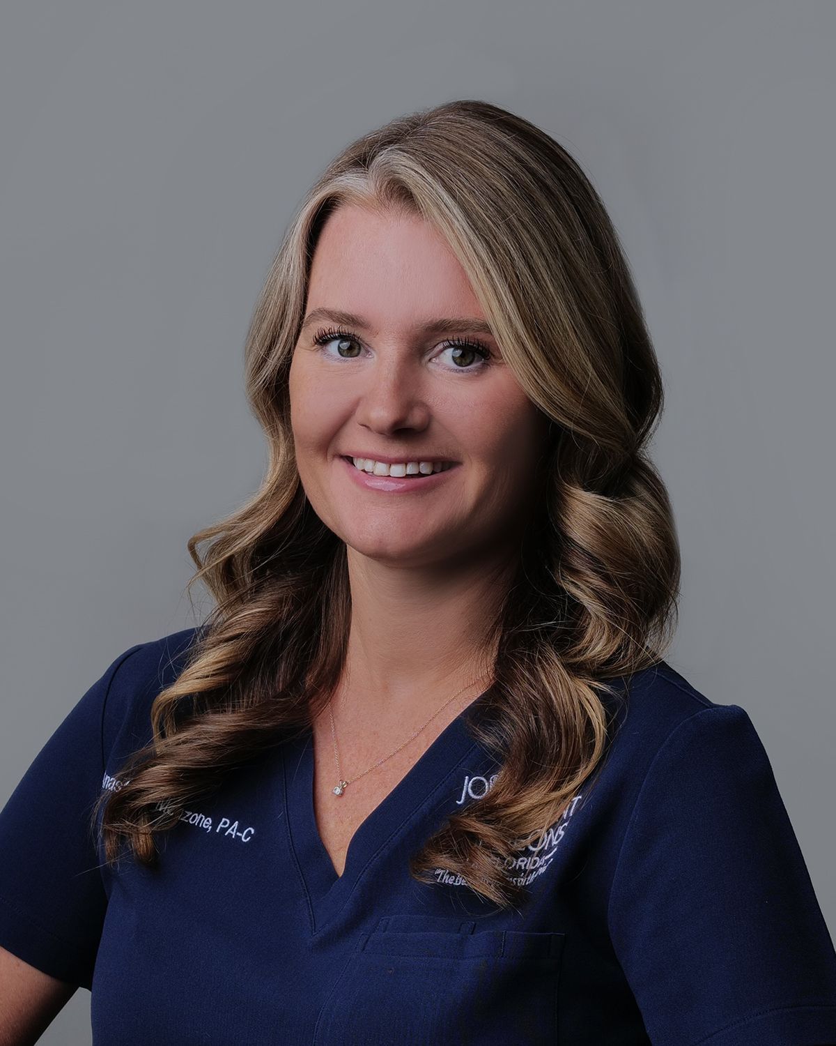 Woman with long blonde hair in a blue scrub top smiles in a medical setting.
