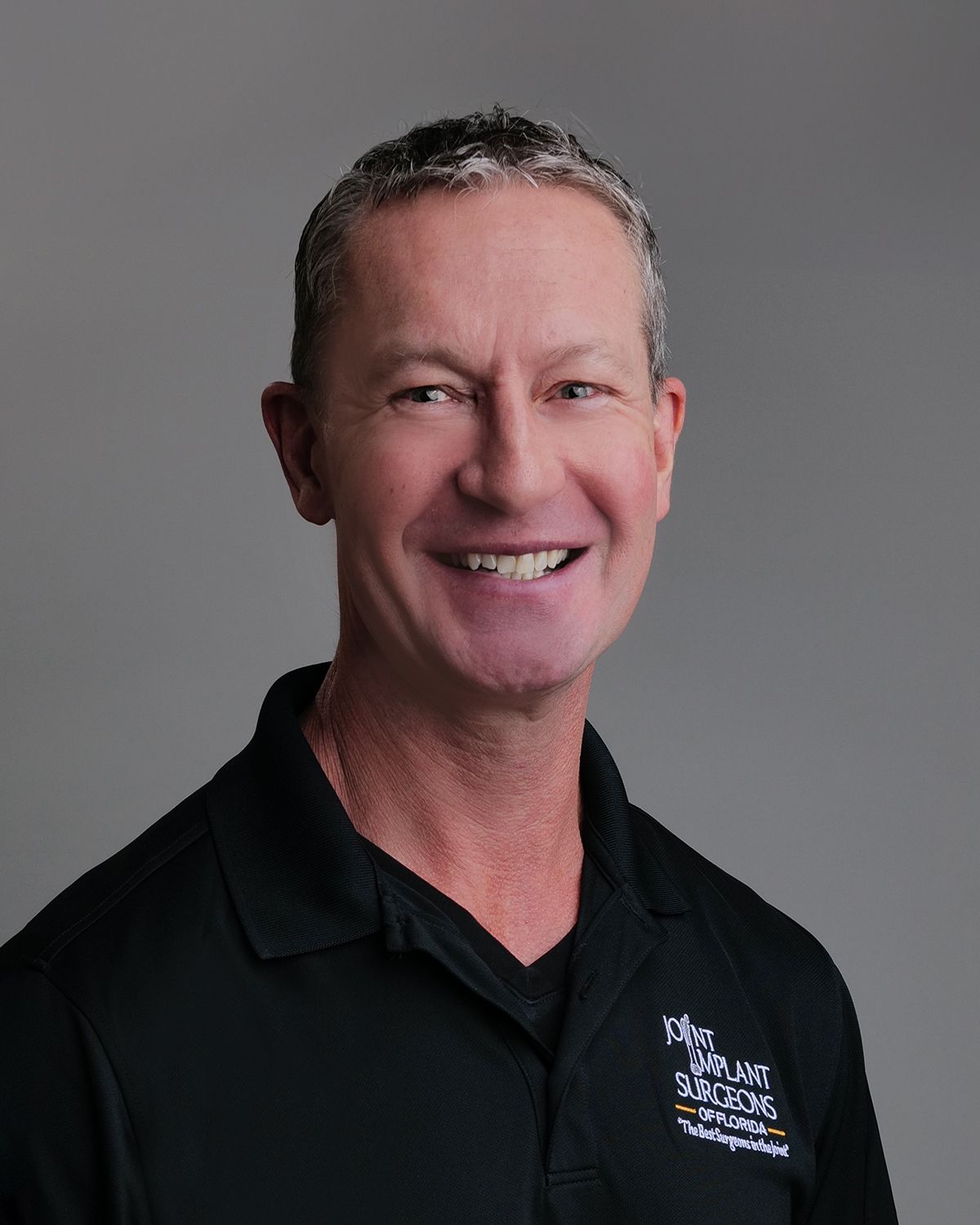 Man in black v-neck shirt smiles in a blurred surgical setting.