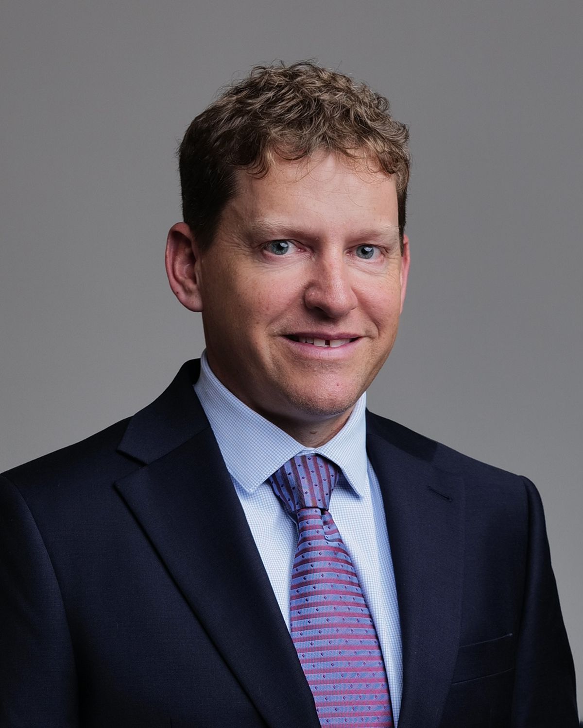 Man in a suit smiles at the camera; background blurred.