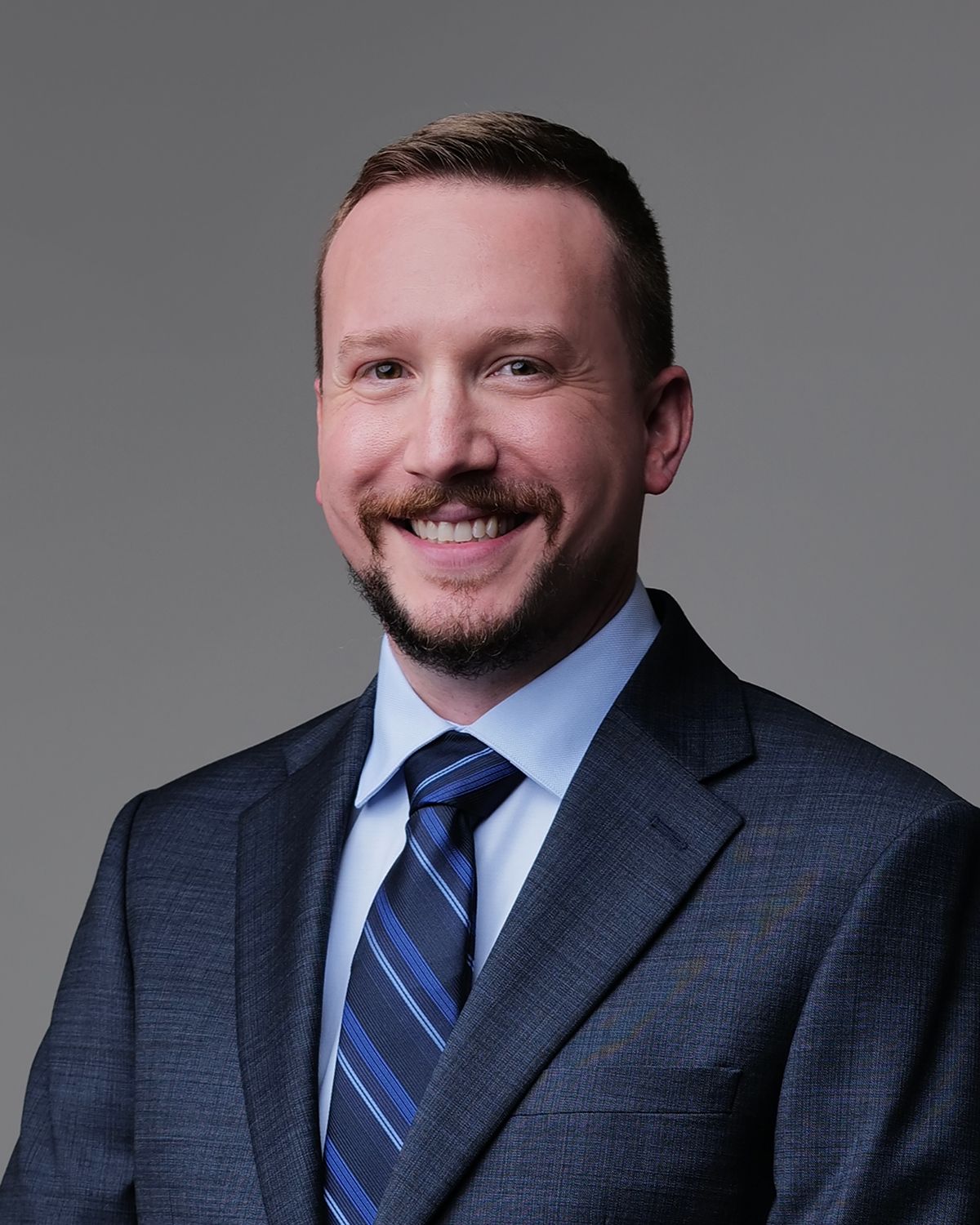 Man in blue suit and tie smiles at the camera, with glasses, in a medical setting.