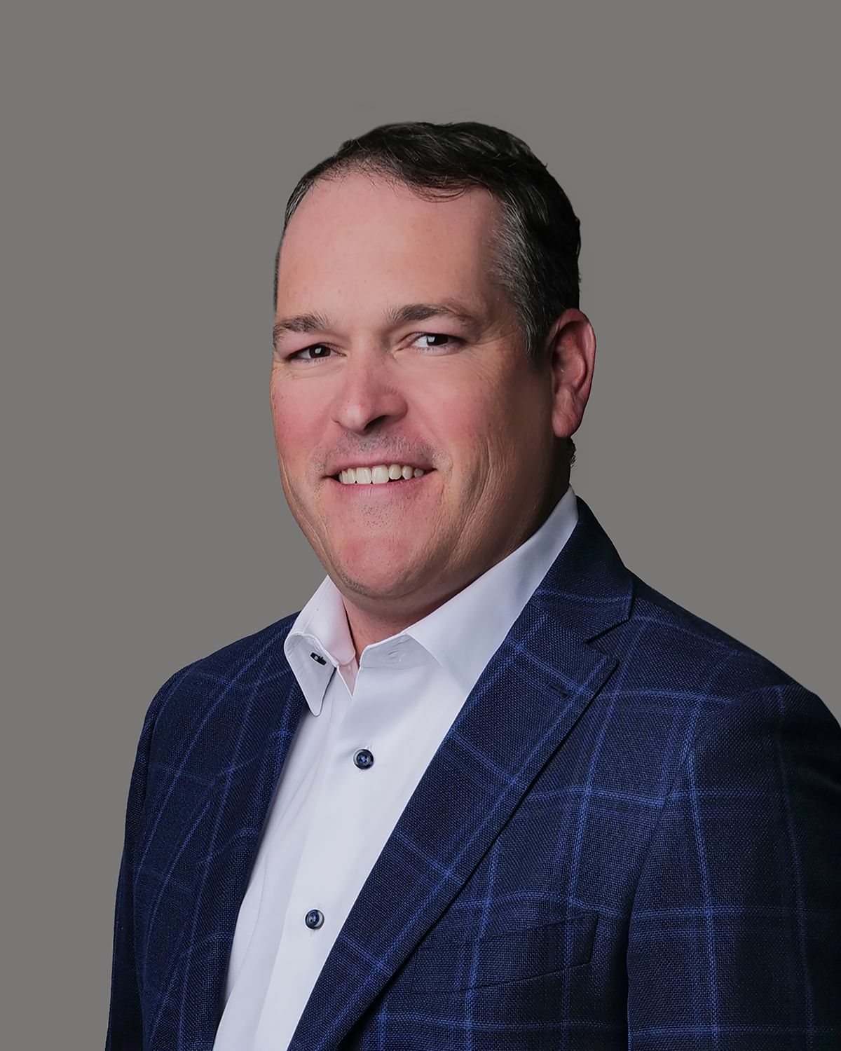 Man in blue plaid suit and tie, smiling, set in an indistinct office environment.