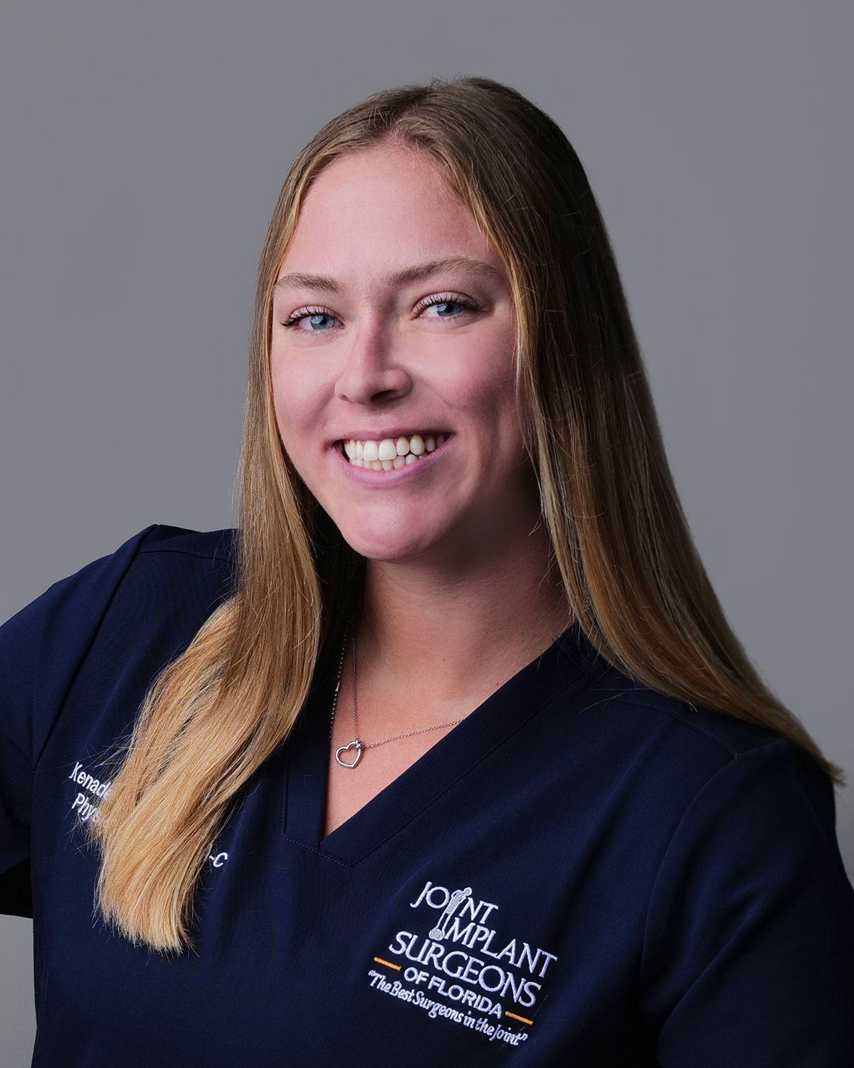 Woman with blonde wavy hair smiles at the camera, wearing a black top, small necklace, in a medical setting.