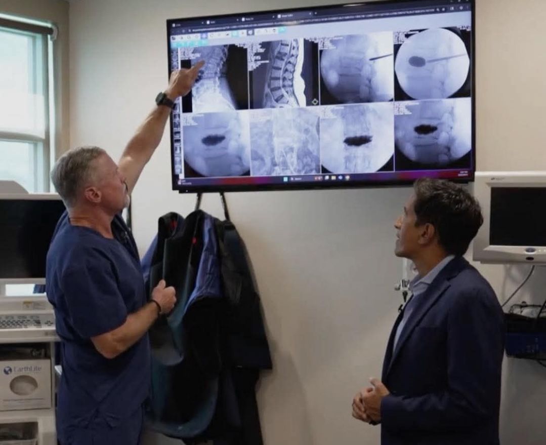 Two men examining spine X-rays on a monitor; one points at the images while the other watches.