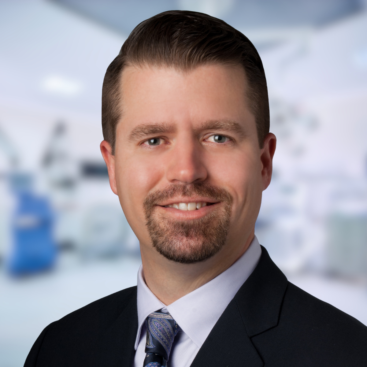 Man in dark suit smiles, with a medical setting blurred in background.