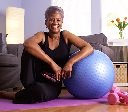 Smiling Black woman rests near exercise ball on a mat; dumbbells nearby.
