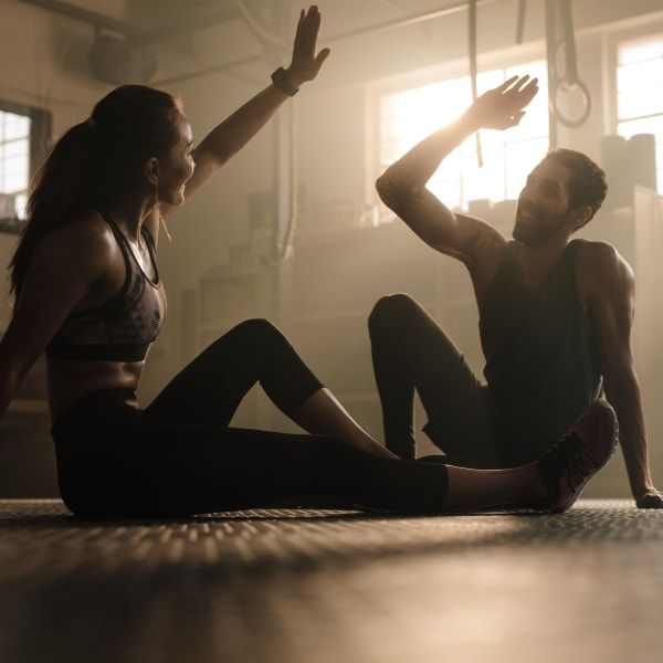 Two people in a gym high-fiving while sitting on the floor after a workout, backlit by bright window light.