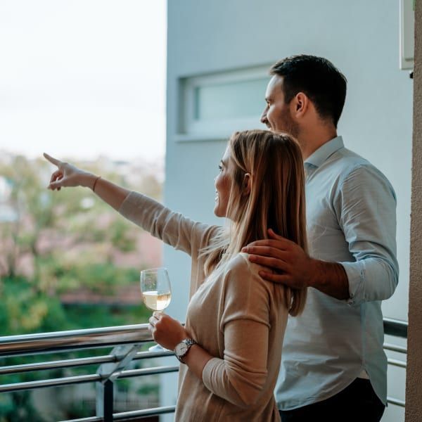 A person holds a wine glass while pointing at a view from a balcony, with another person&rsquo;s hand resting on their shoulder.