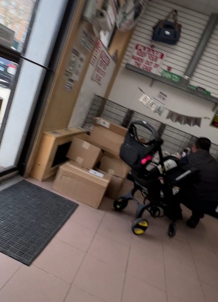 A person crouches near a stroller inside a store. Cardboard boxes are stacked near the entrance.