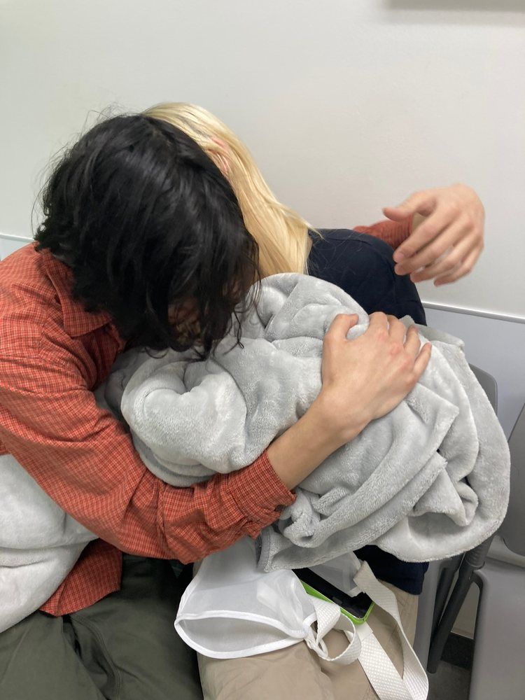 Two people embracing, holding a gray blanket. One has black hair, the other blonde. Indoors.