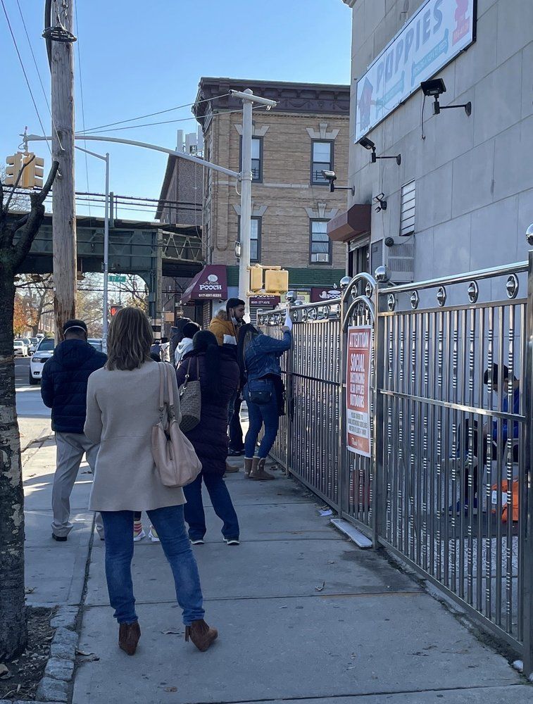 People line up outside a business, with a metal fence and overhead train tracks visible.