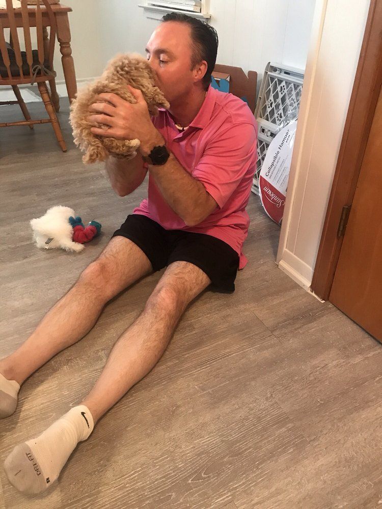 Man in pink shirt kneels, kissing a fluffy, tan puppy. Sitting on a wood floor.