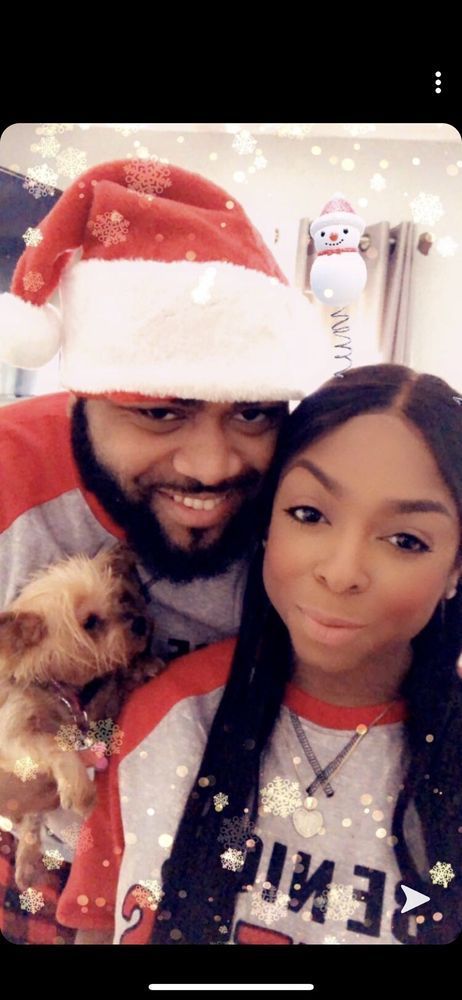 A couple with a dog pose for a Christmas photo. The man wears a Santa hat. Snowflakes fall.