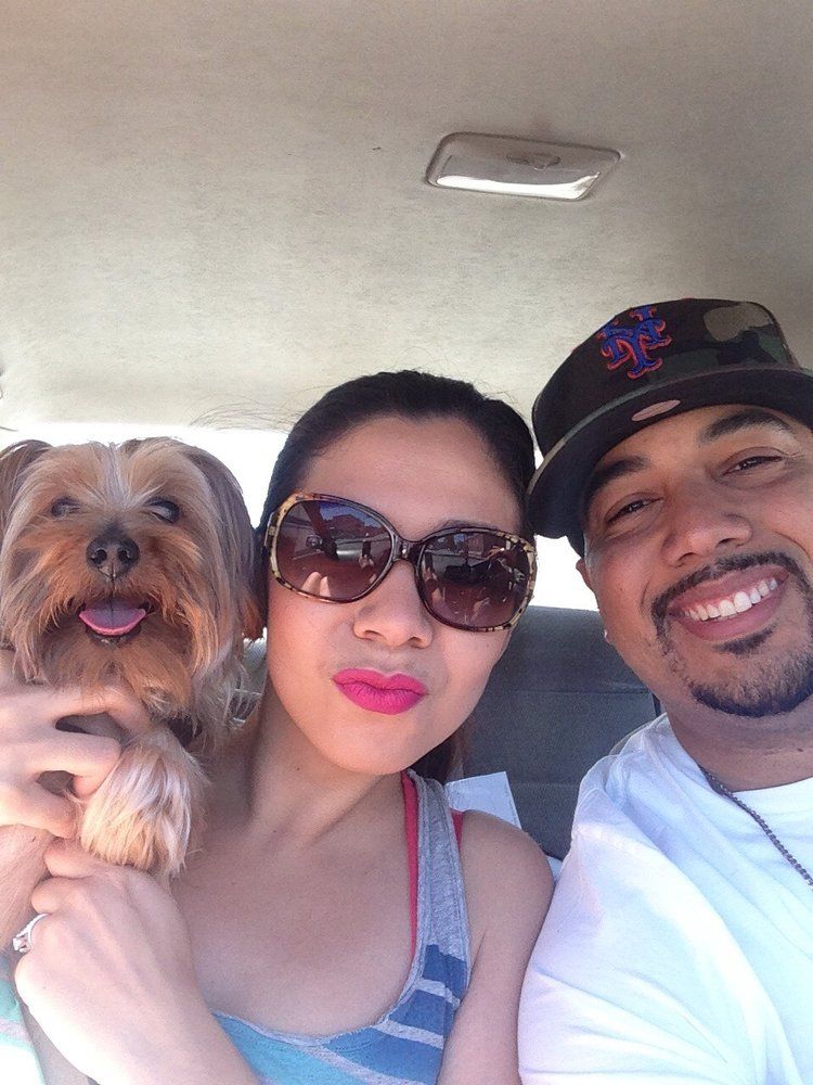 Person holding dog smiles, joined by person with hat, selfie inside a car.