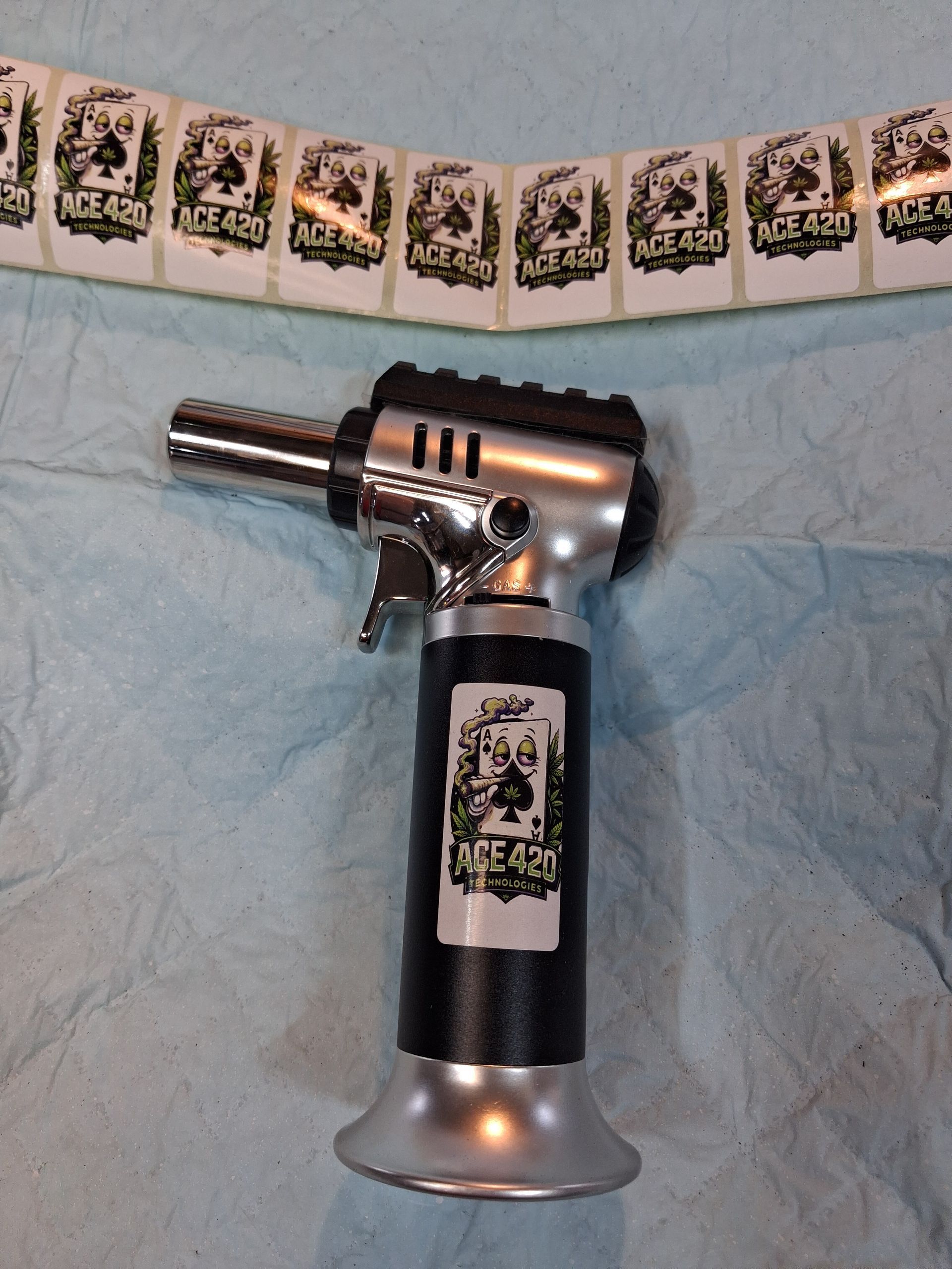 Silver handheld butane torch on a blue cloth, with a strip of small black-and-white labels above it