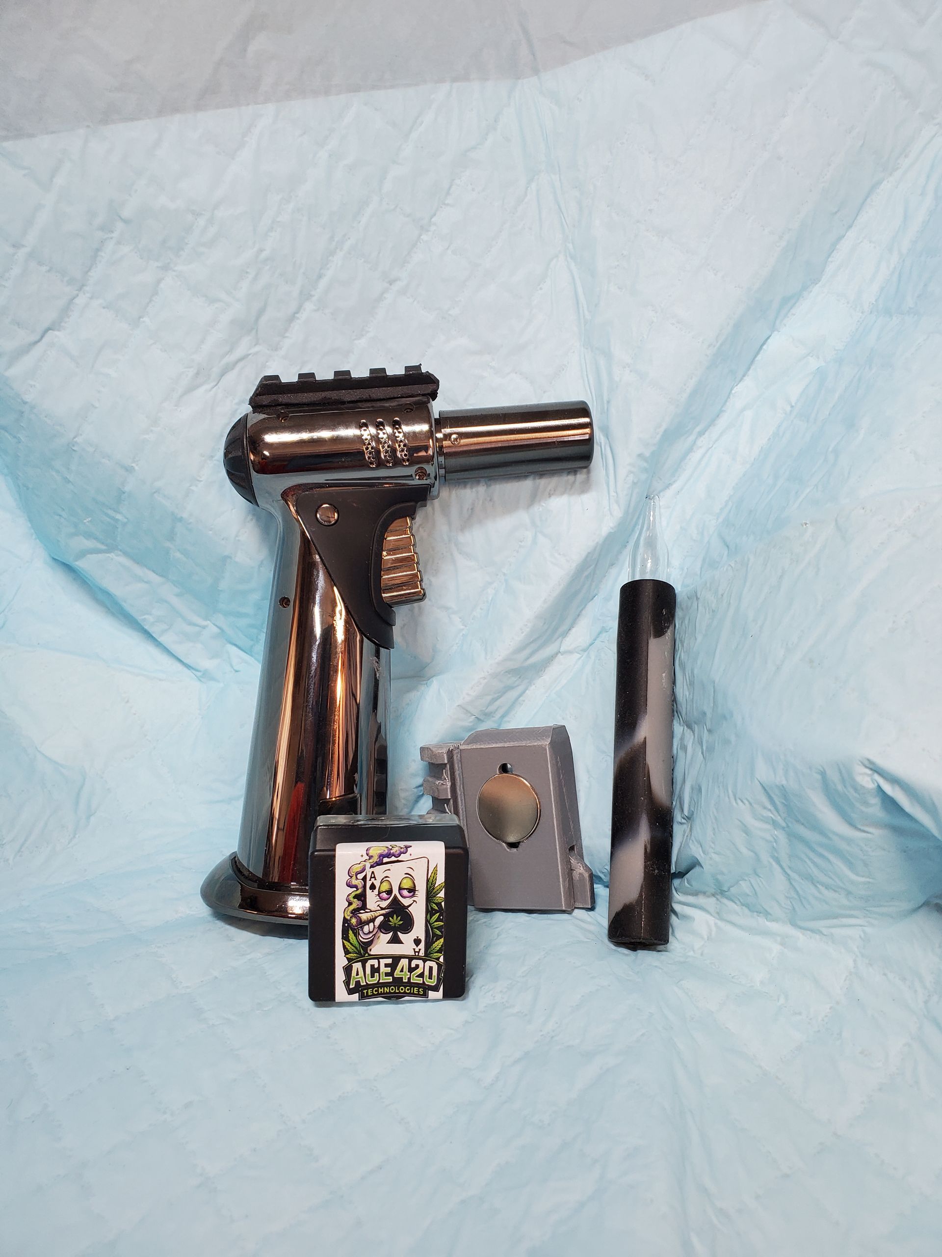 Small black and silver handheld torch lighter, a metal piece, and a black cylindrical accessory on blue fabric.