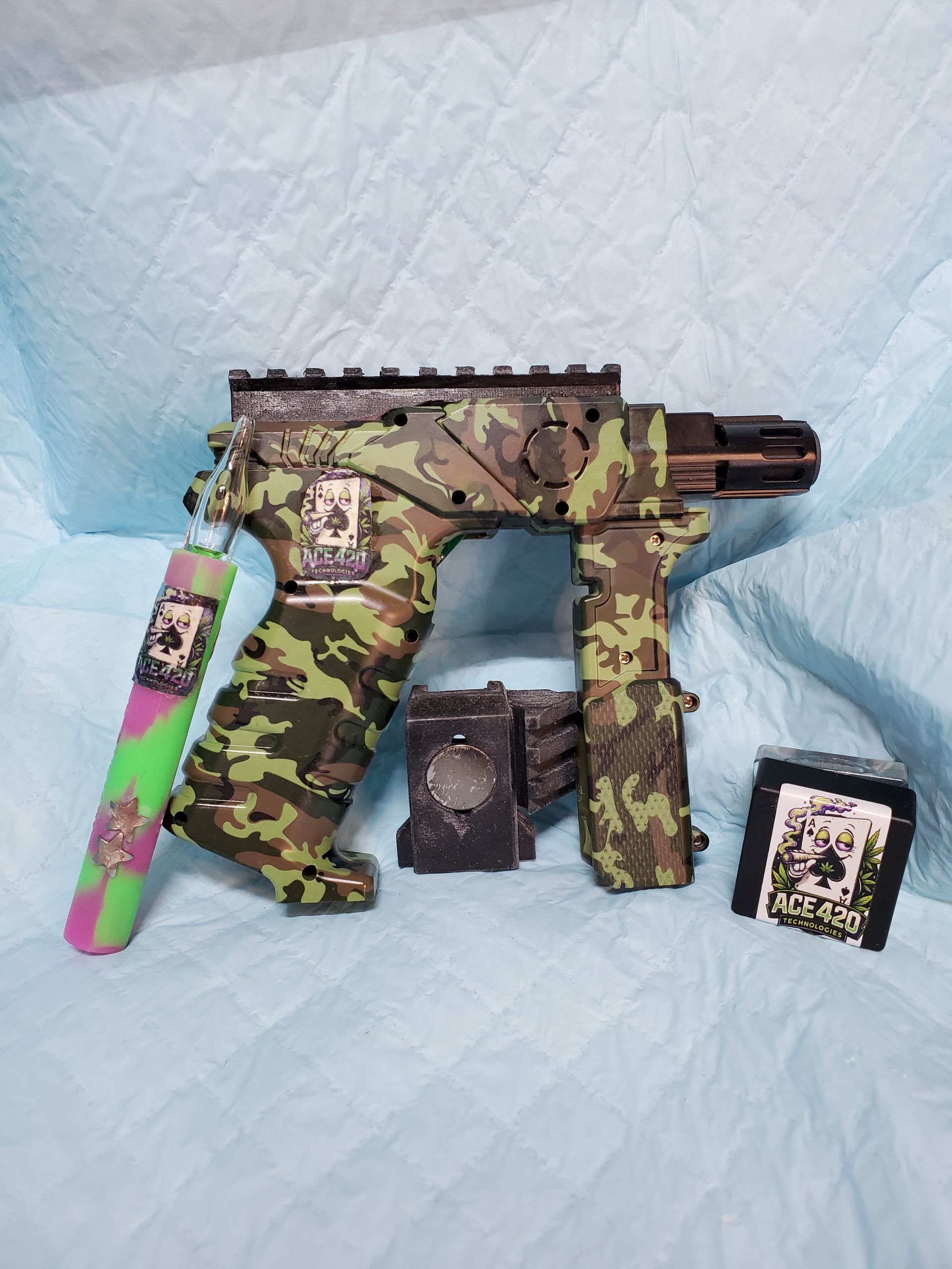 Camouflage toy submachine gun with pink and green magazine, shown with two small accessories on a blue background