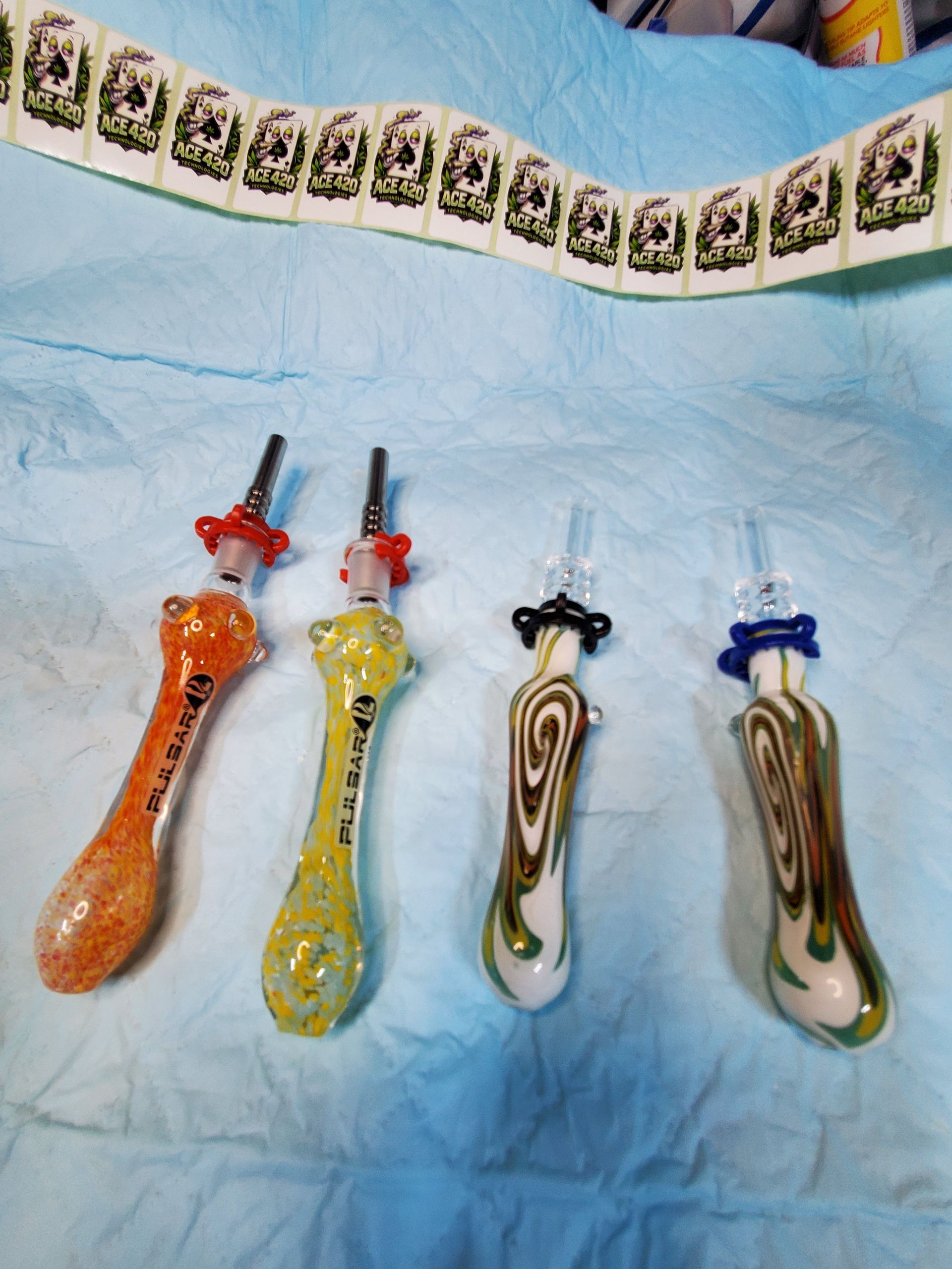 Four colorful glass smoking pipes on a blue cloth, with orange, yellow, and patterned green-white stems and bowls
