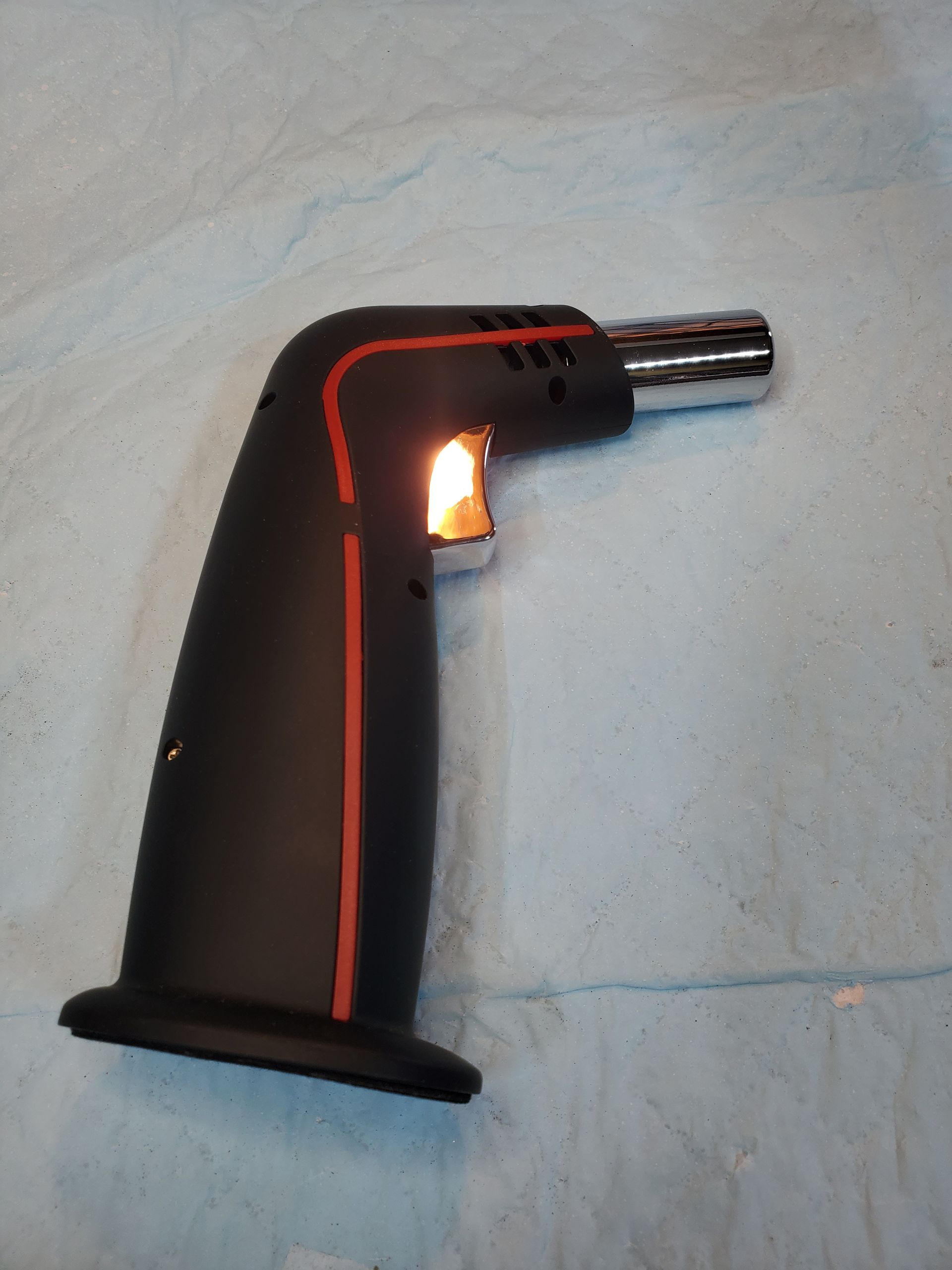 Black handheld butane torch with orange flame on a light cloth background