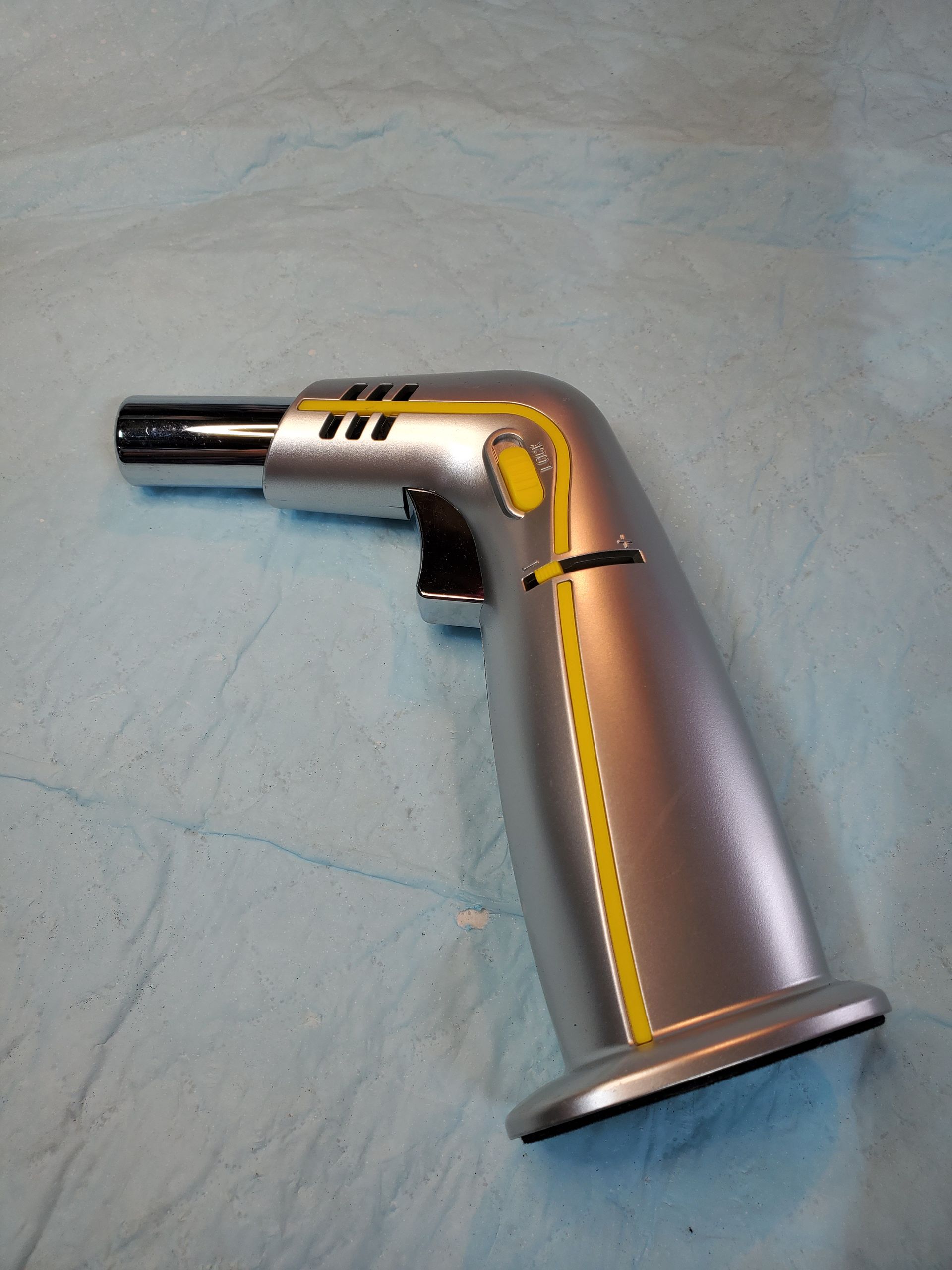 Metal handheld torch lighter with silver body and black nozzle on a light blue surface