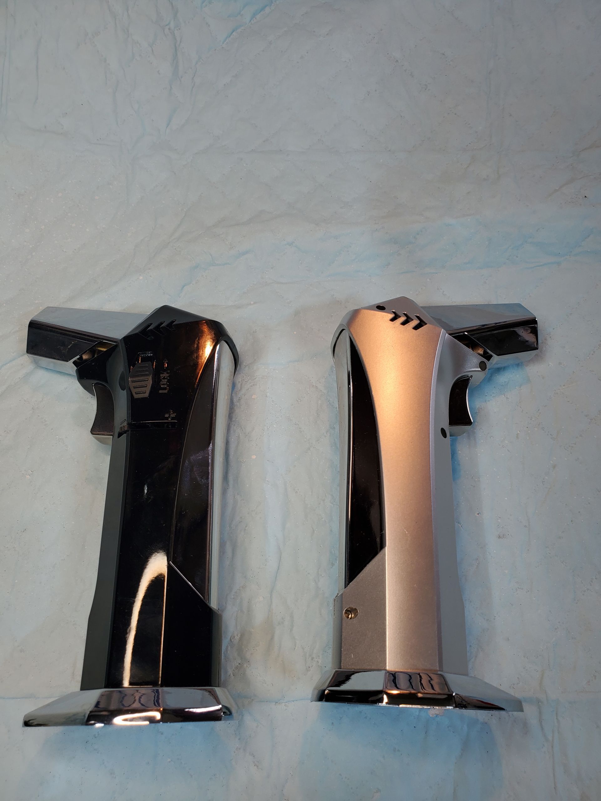 Two chrome T-shaped faucet handles on a light countertop, one dark and one reflective silver.