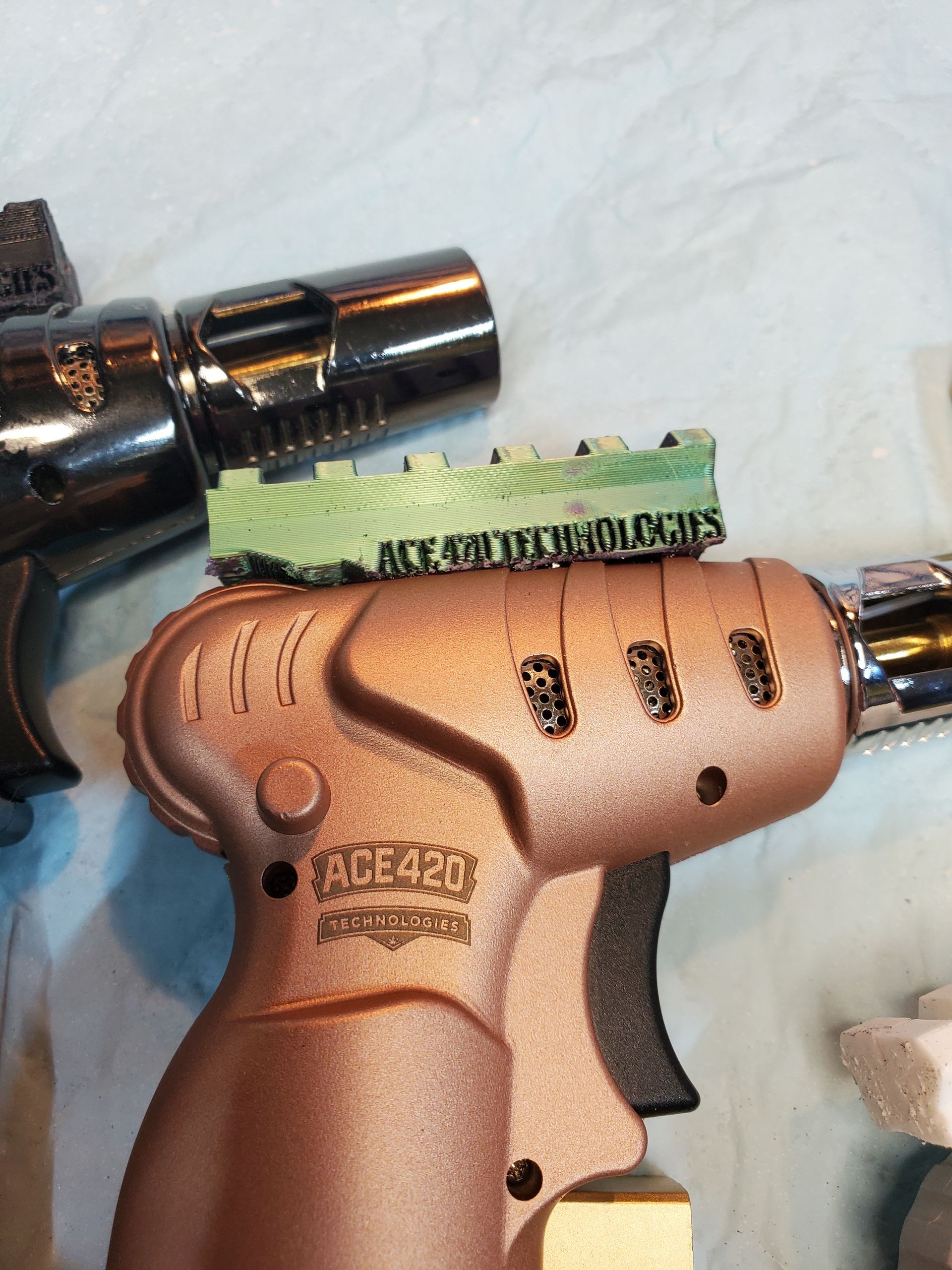 Copper-colored handgun with green putty on top and a metal barrel attachment, on a white cloth background