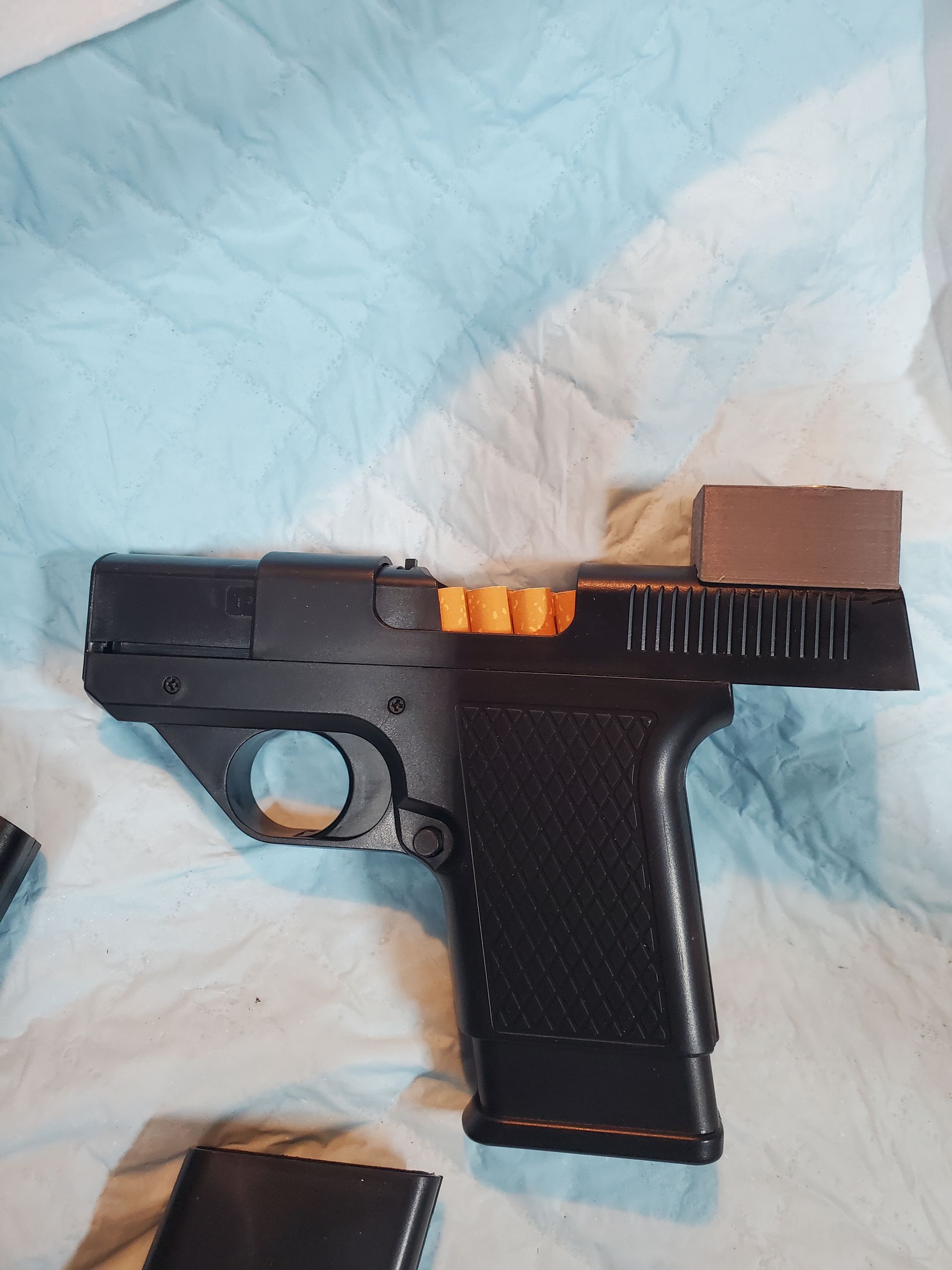 Black handgun with orange chamber cover and brown tape on a light blue bedspread.