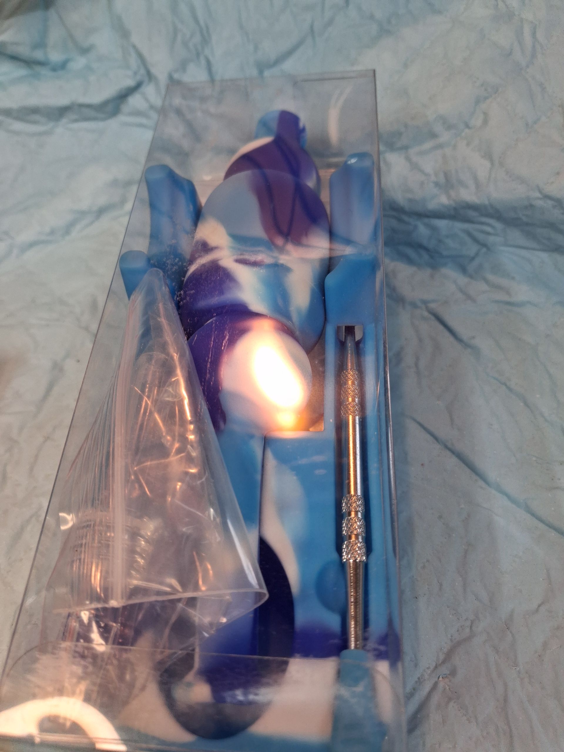 Clear plastic package with a blue and purple handheld item and a small silver metal tool on a wrinkled blue cloth.