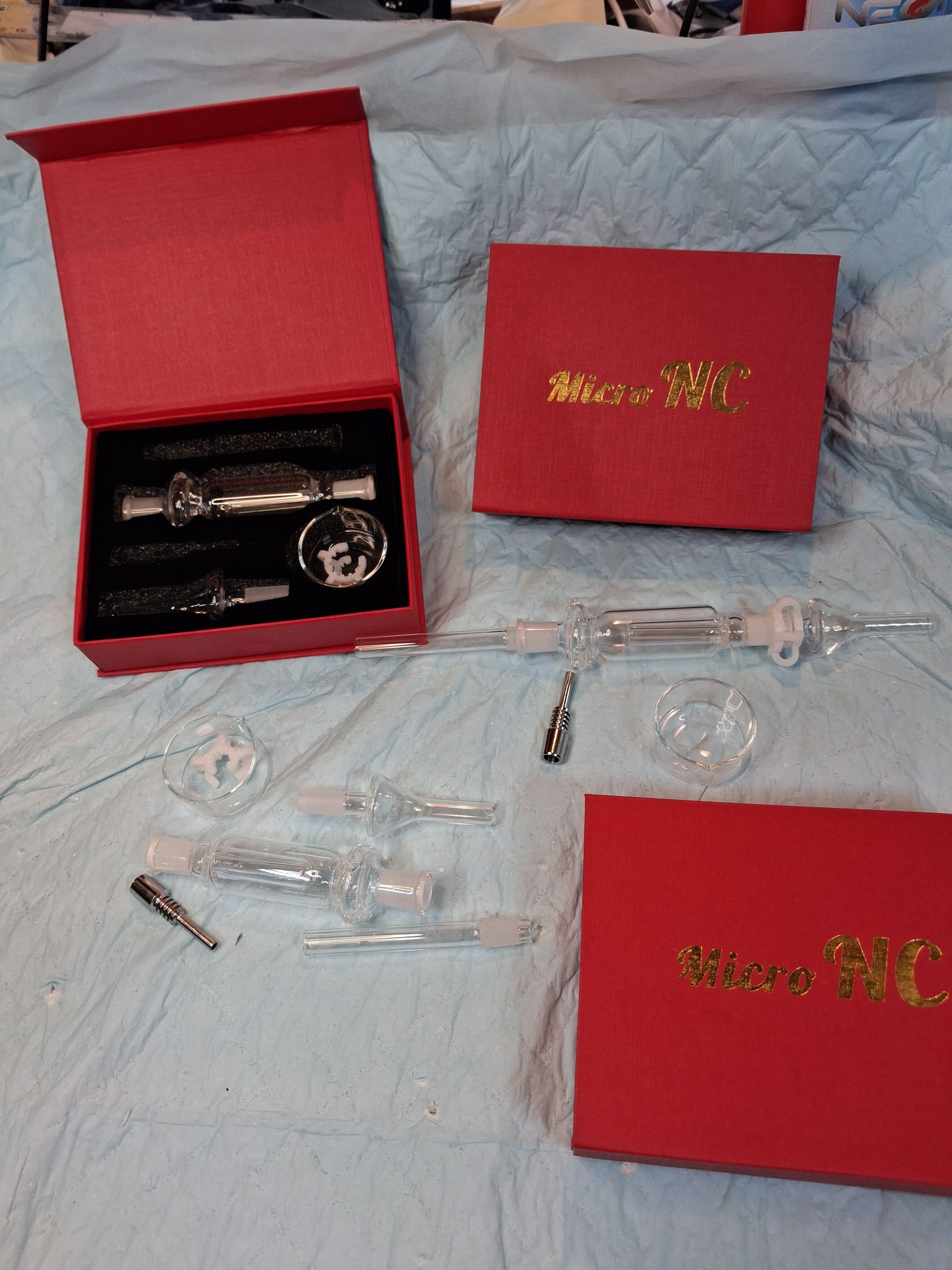 Red gift boxes with gold lettering and several small clear and black packaged items on a bedspread