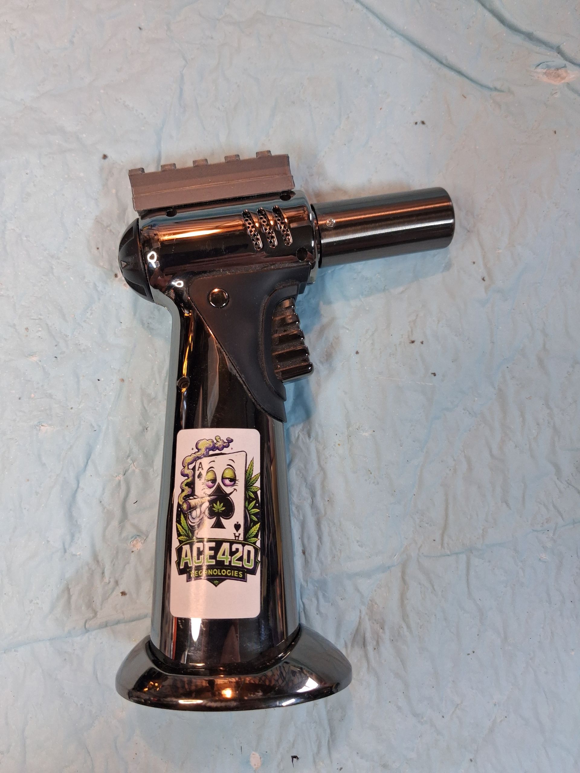 Black torch lighter with a long nozzle standing upright against a light blue wall.