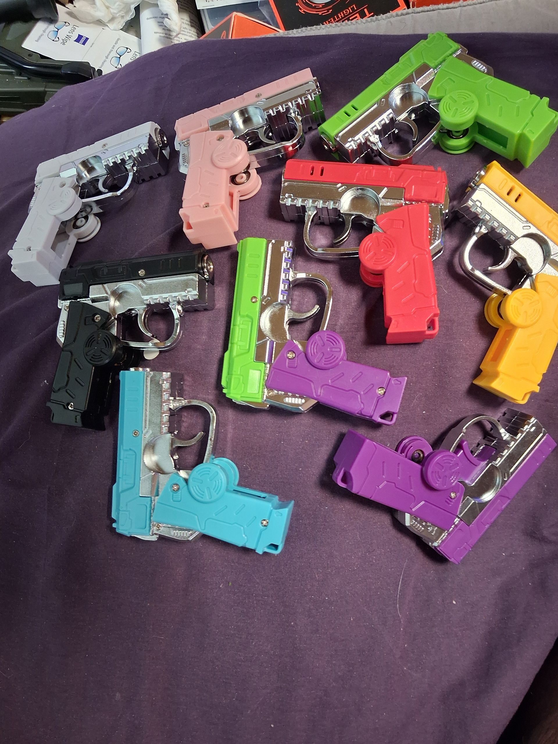 Assorted colorful gun shaped foldable butane torch lighters/fidget spinners, scattered on a purple fabric background