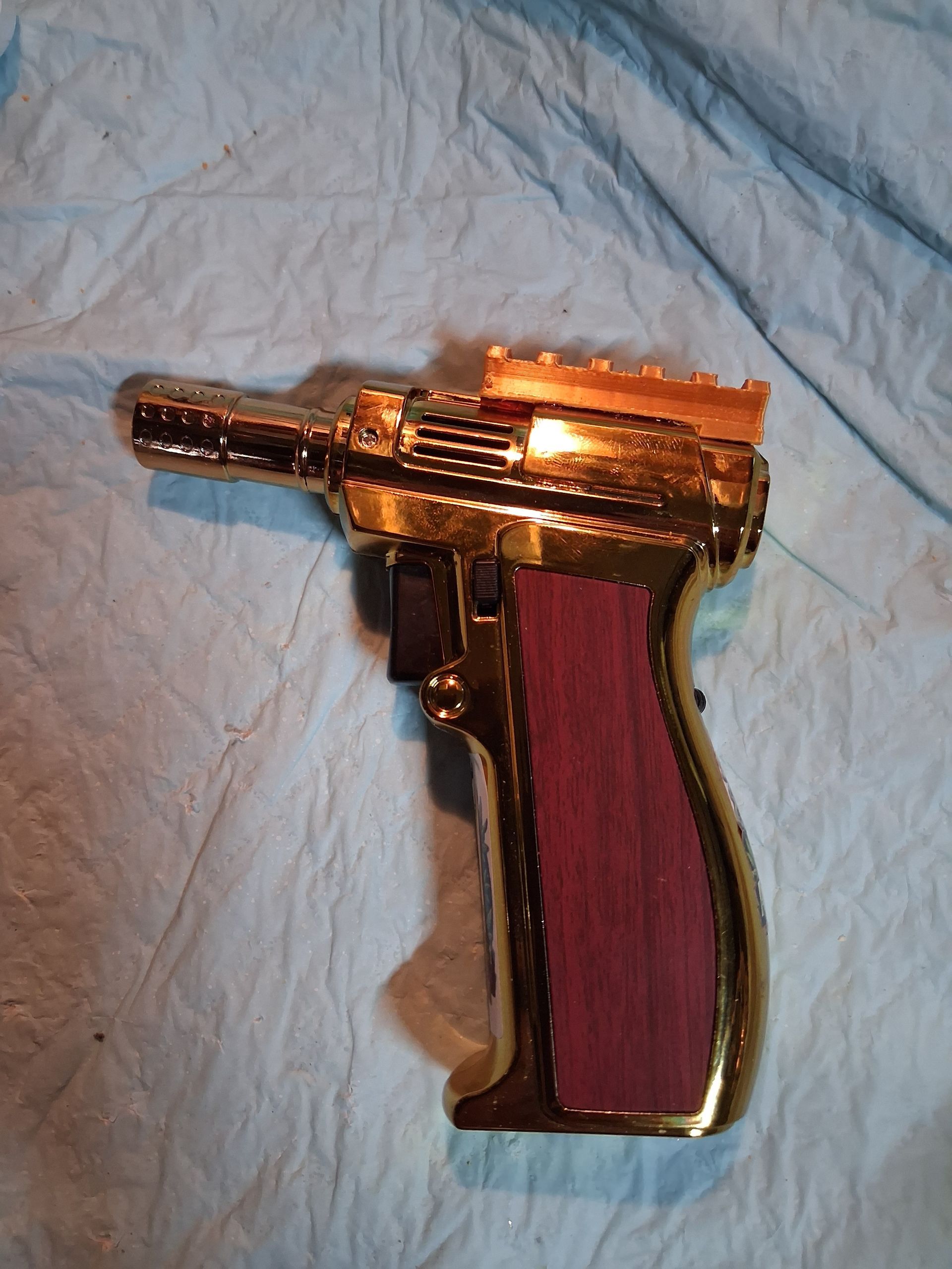 Gold-colored gun shaped butane torch with a faux wood grip on a wrinkled light blue cloth background