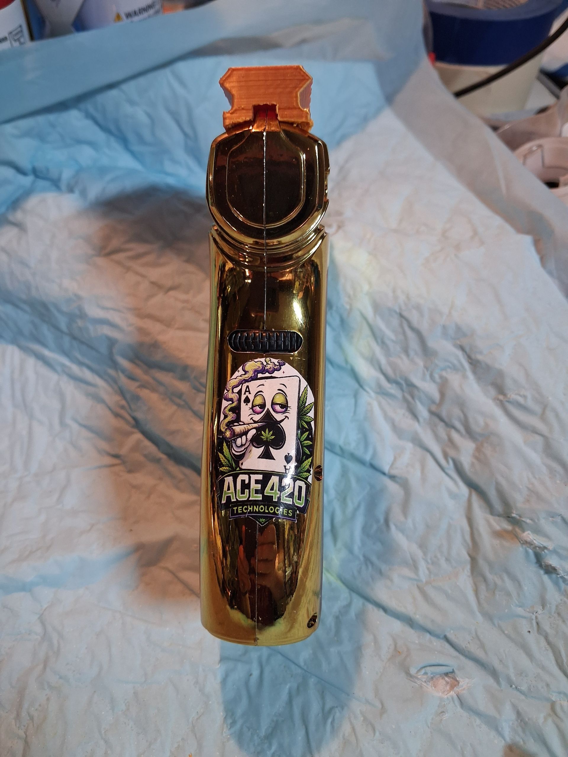 Gold torch lighter standing with a logo sticker label and the word “ACE420 Technologies” on a blue cloth.
