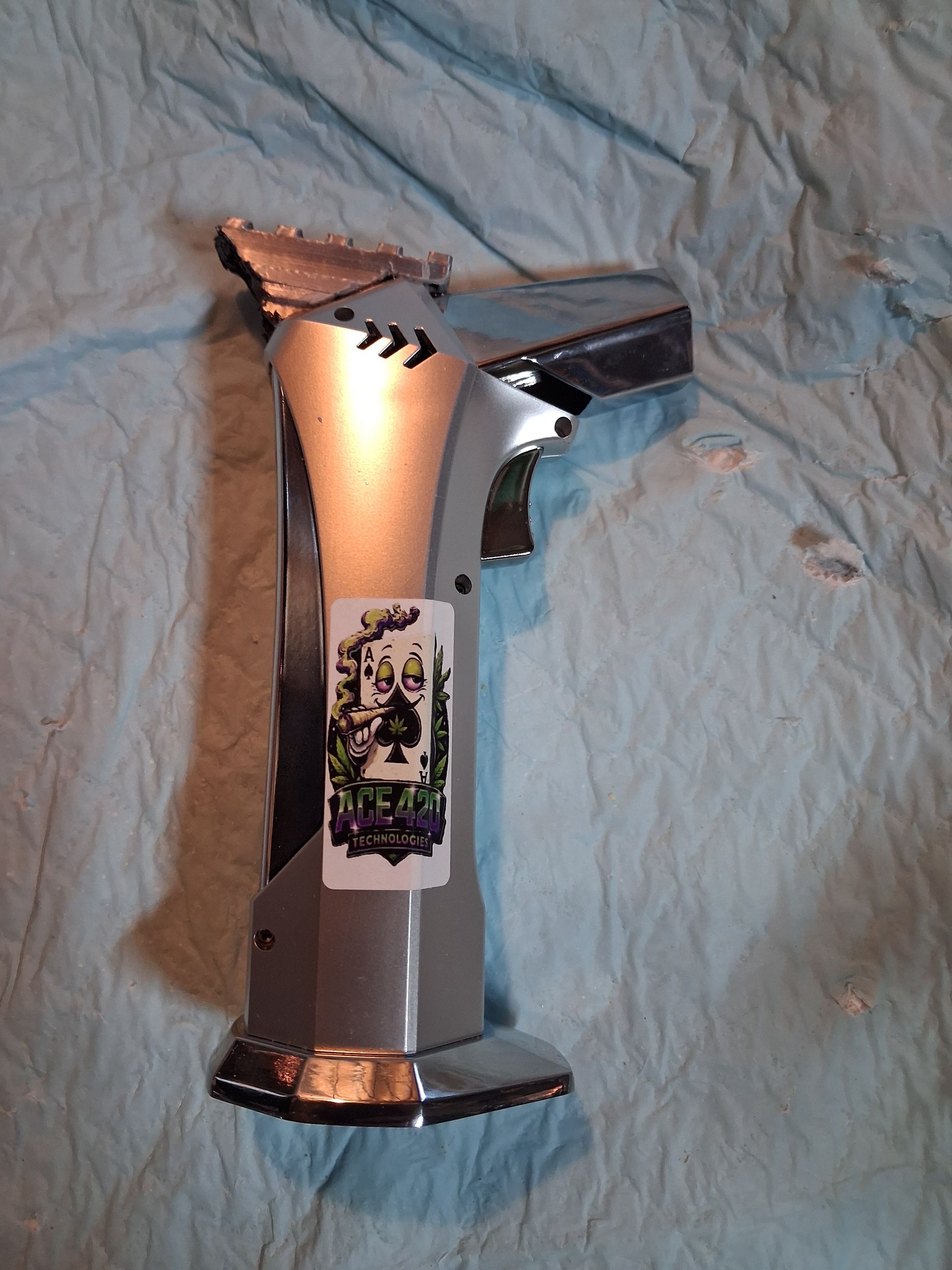 Metallic silver handheld butane torch lying on a wrinkled light blue fabric surface.