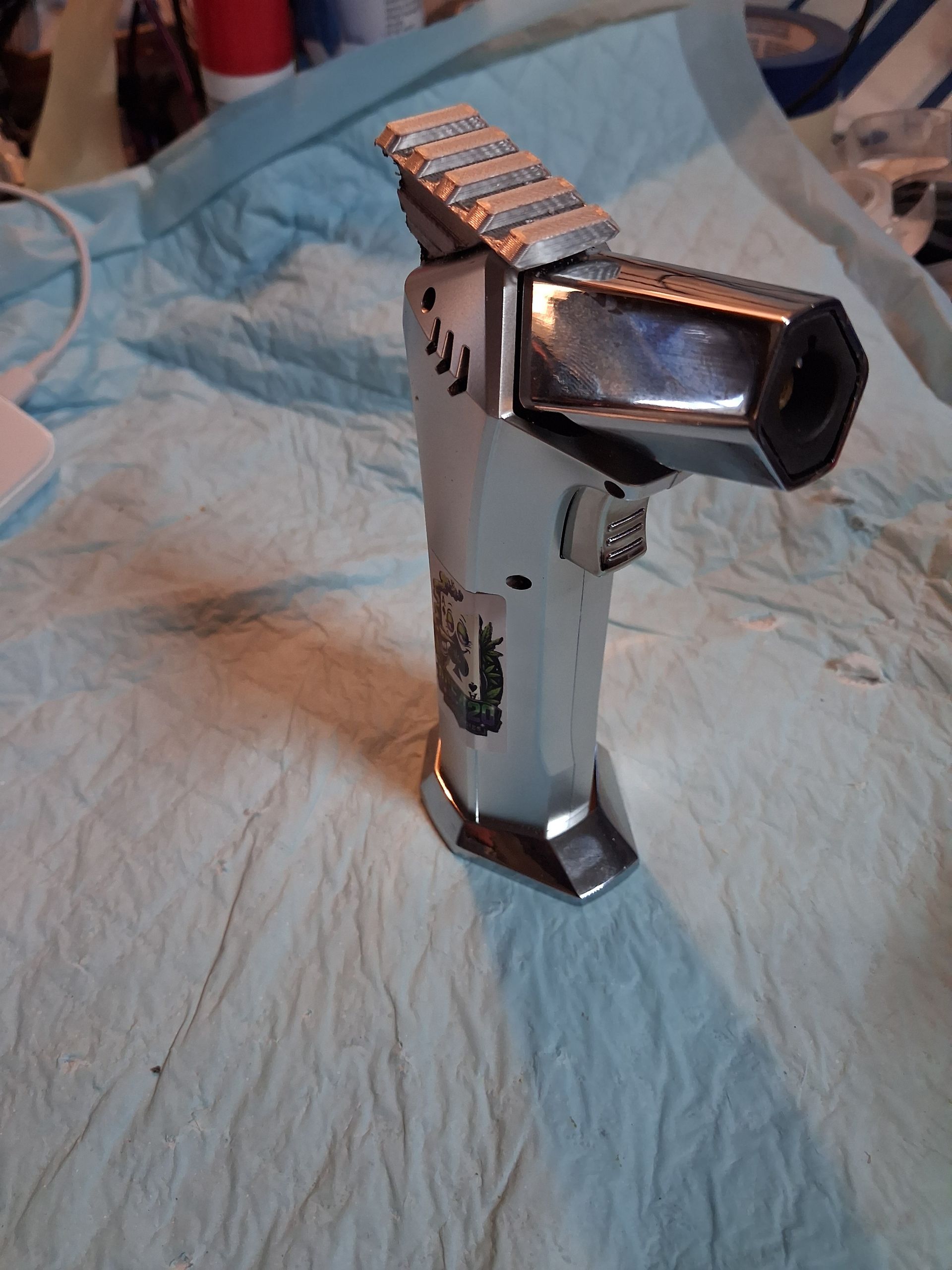Small silver metallic Butane torch device with a rail on top, standing on a light blue cloth.