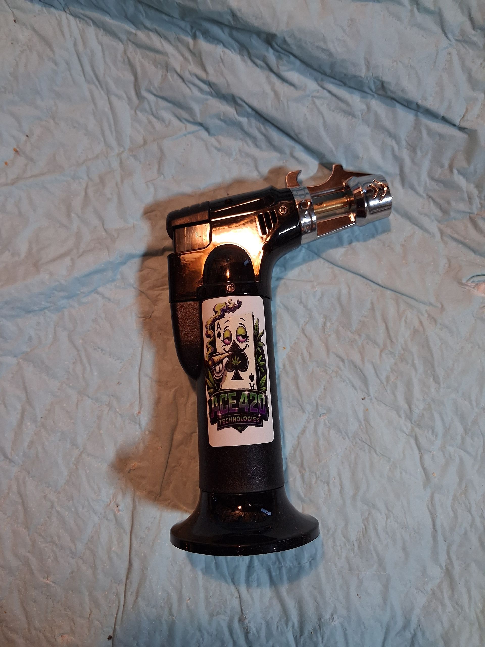 Chrome torch lighter with a black handle and sticker, lying on a wrinkled light blue cloth.