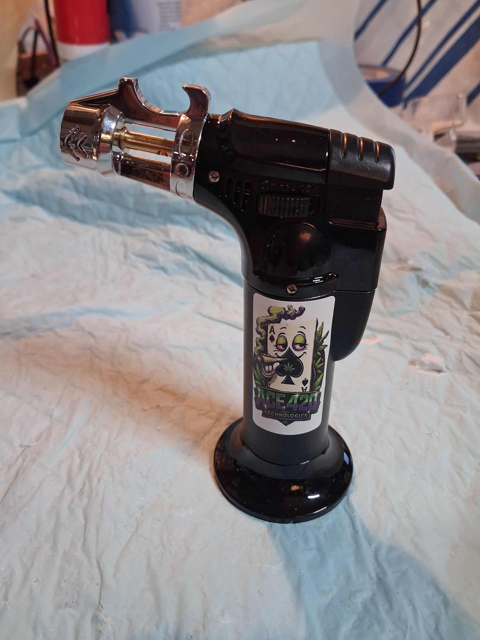 Black-and-silver metallic butane torch lighter with a bottle opener on a light fabric surface