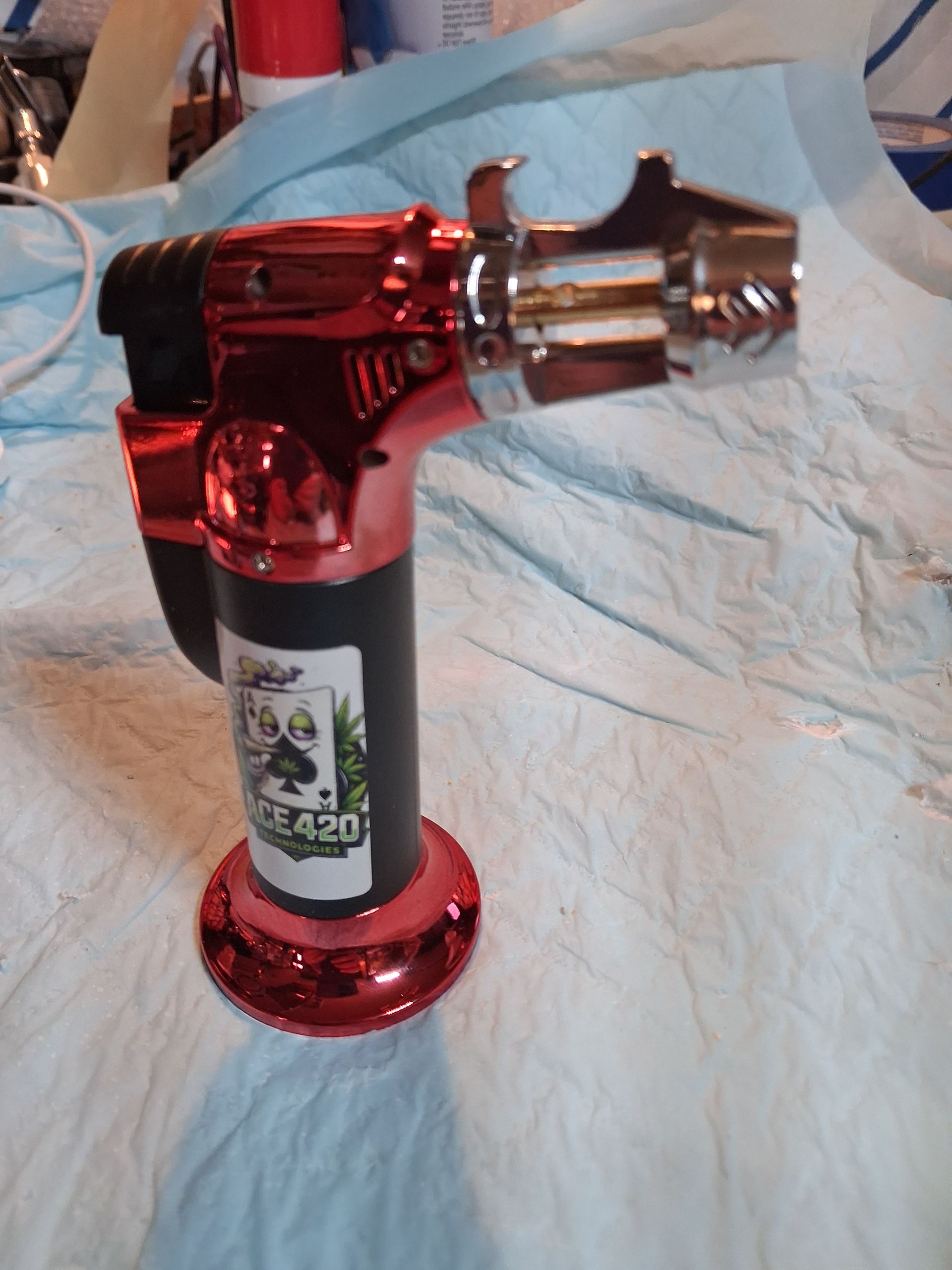 Red metallic butane torch lighter with a bottle opener on a light fabric surface.