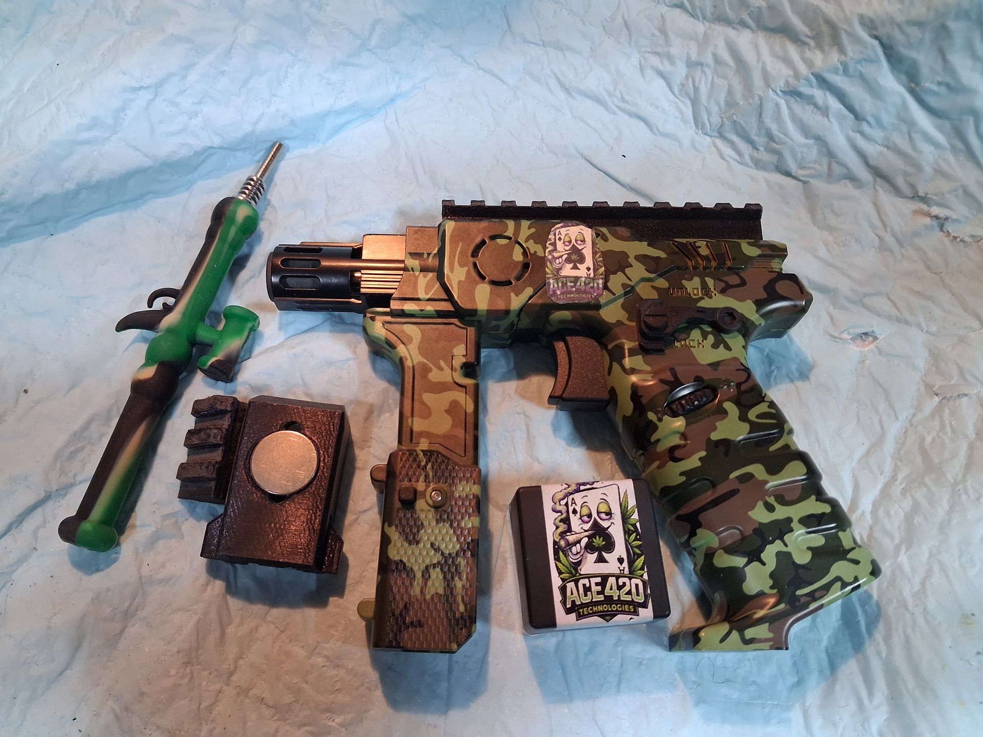Toy paintball-style gun with green camouflage grip, tank, pouch, and cards on a blue cloth background